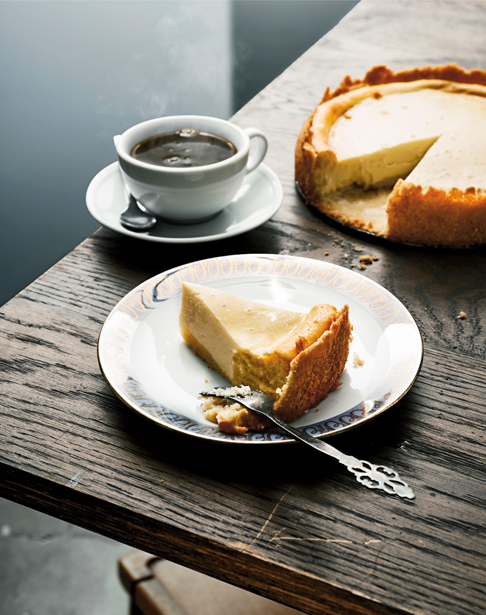 En bit cheesecake ligger på en vit tallrik på ett rustikt träbord. Bredvid står en kopp svart kaffe på ett fat. I bakgrunden syns en större cheesecake med en bit bortskuren. Ljuset är mjukt och naturligt, och framhäver den krämiga fyllningen och den gyllene kanten på kakan.