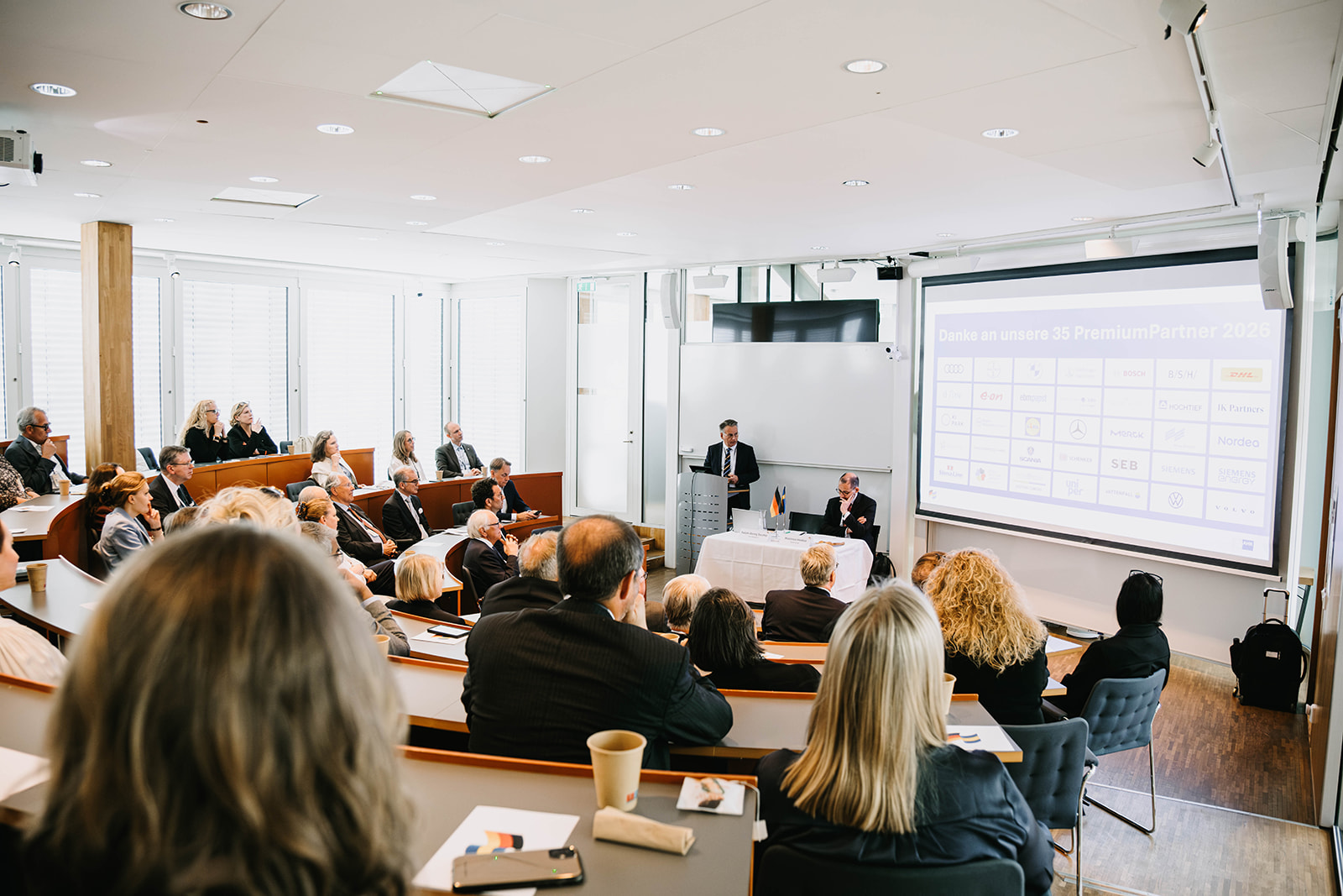 Årsmöte arrangerat av Tysk‑Svenska Handelskammaren i en föreläsningssal på Handelshögskolan i Stockholm. Deltagare sitter i nivåindelade rader och lyssnar på en presentation längst fram, där en person presenterar innehåll på en stor projicerad skärm.