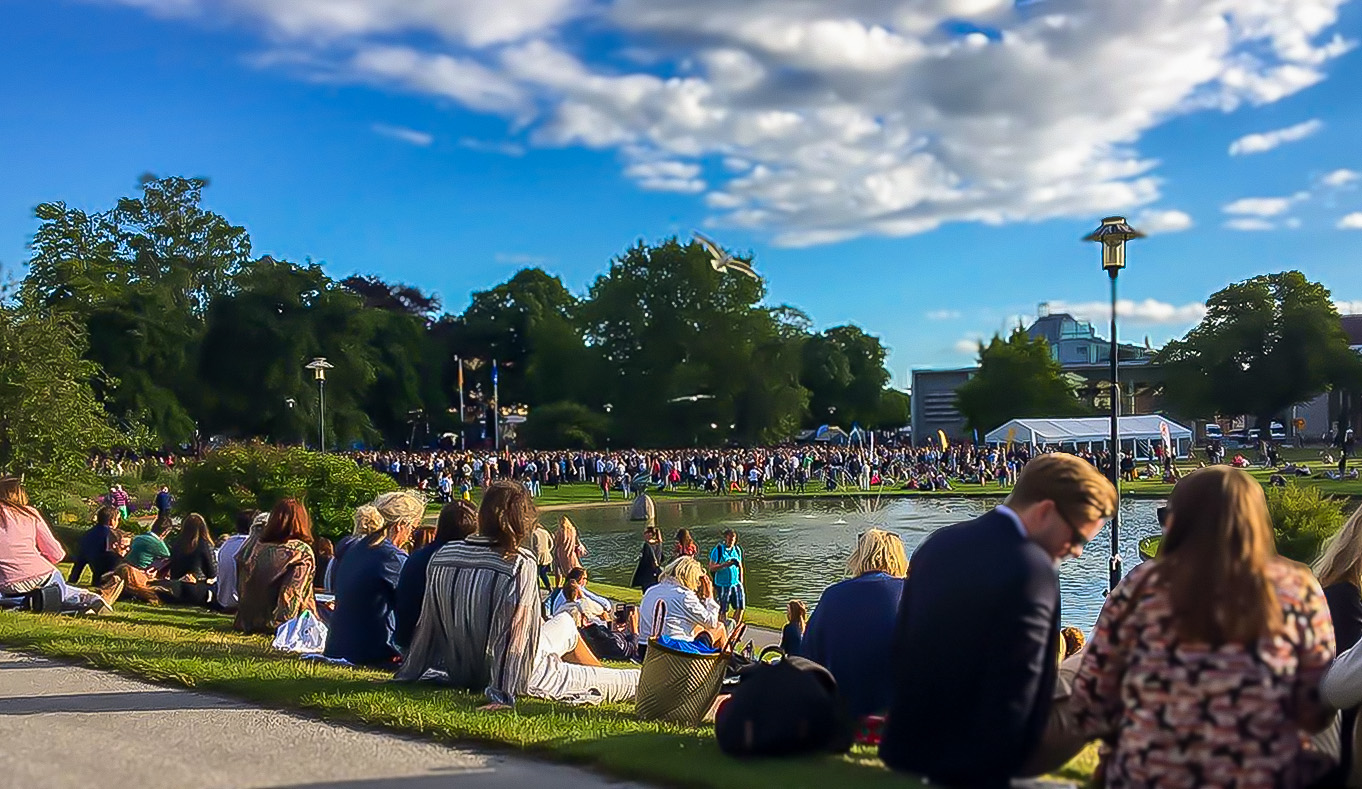 Personer sitter på en gräsmatta vid en damm i en park i Almedalen, i bakgrunden syns en stor folkmassa och träd under en blå himmel med moln.