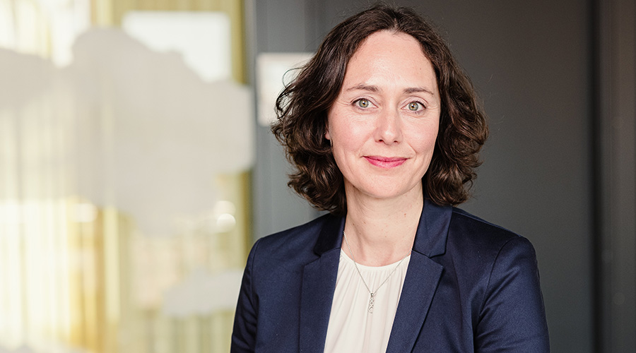 En person klädd i en mörk blazer och ljus tröja står inomhus framför en bakgrund med glaspartier och gardiner.