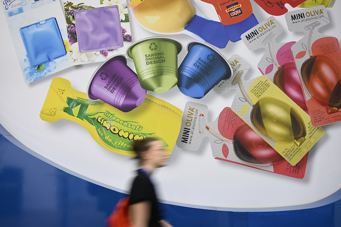 En färgstark vägg med olika kosmetik- och skönhetsförpackningar i plast och folie, medan en förbipasserande person rör sig i förgrunden.