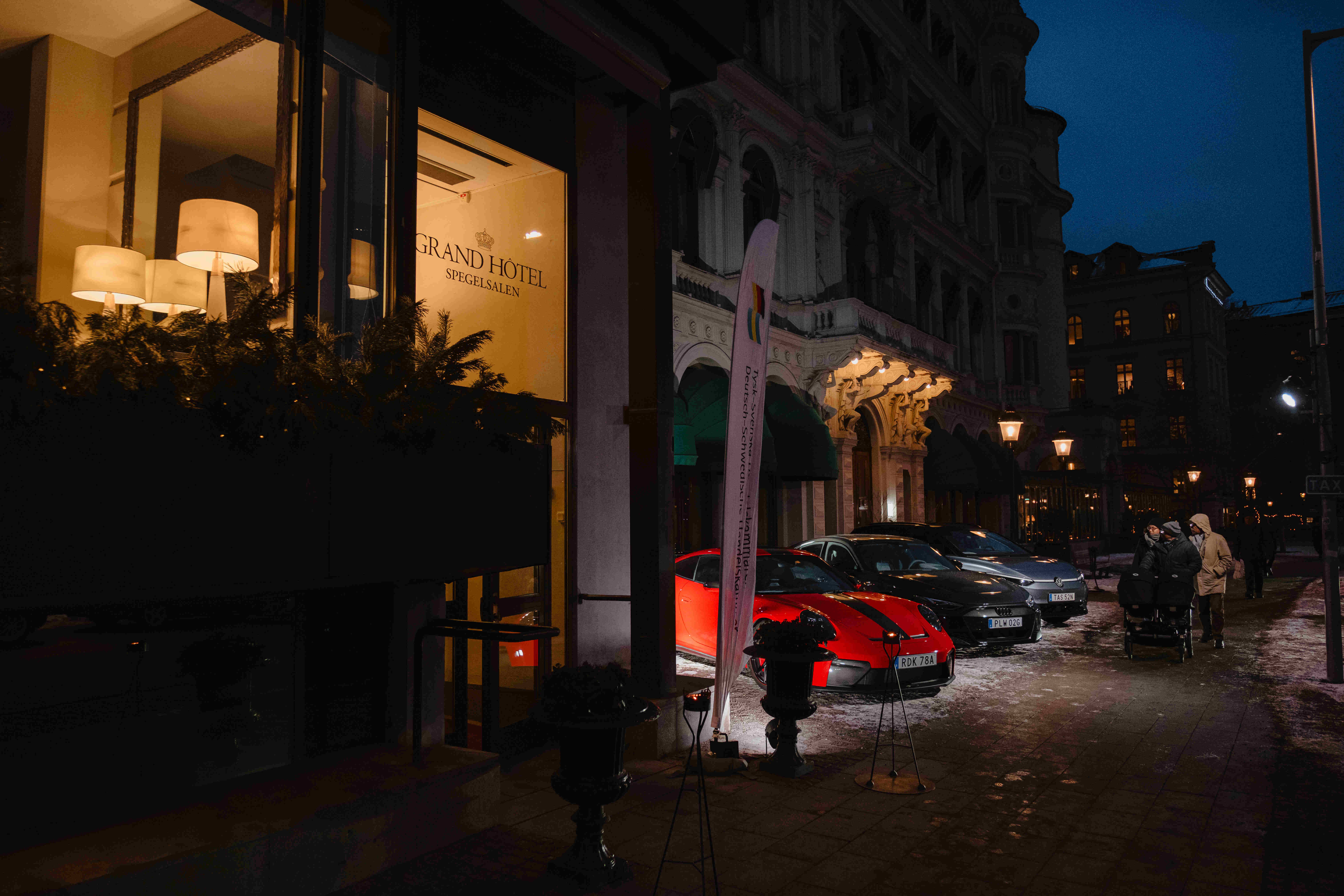 Entréområdet till Grand Hôtel i kvällsbelysning med parkerade bilar, en upplyst flaggskylt och fotgängare längs den vinterbelagda gatan