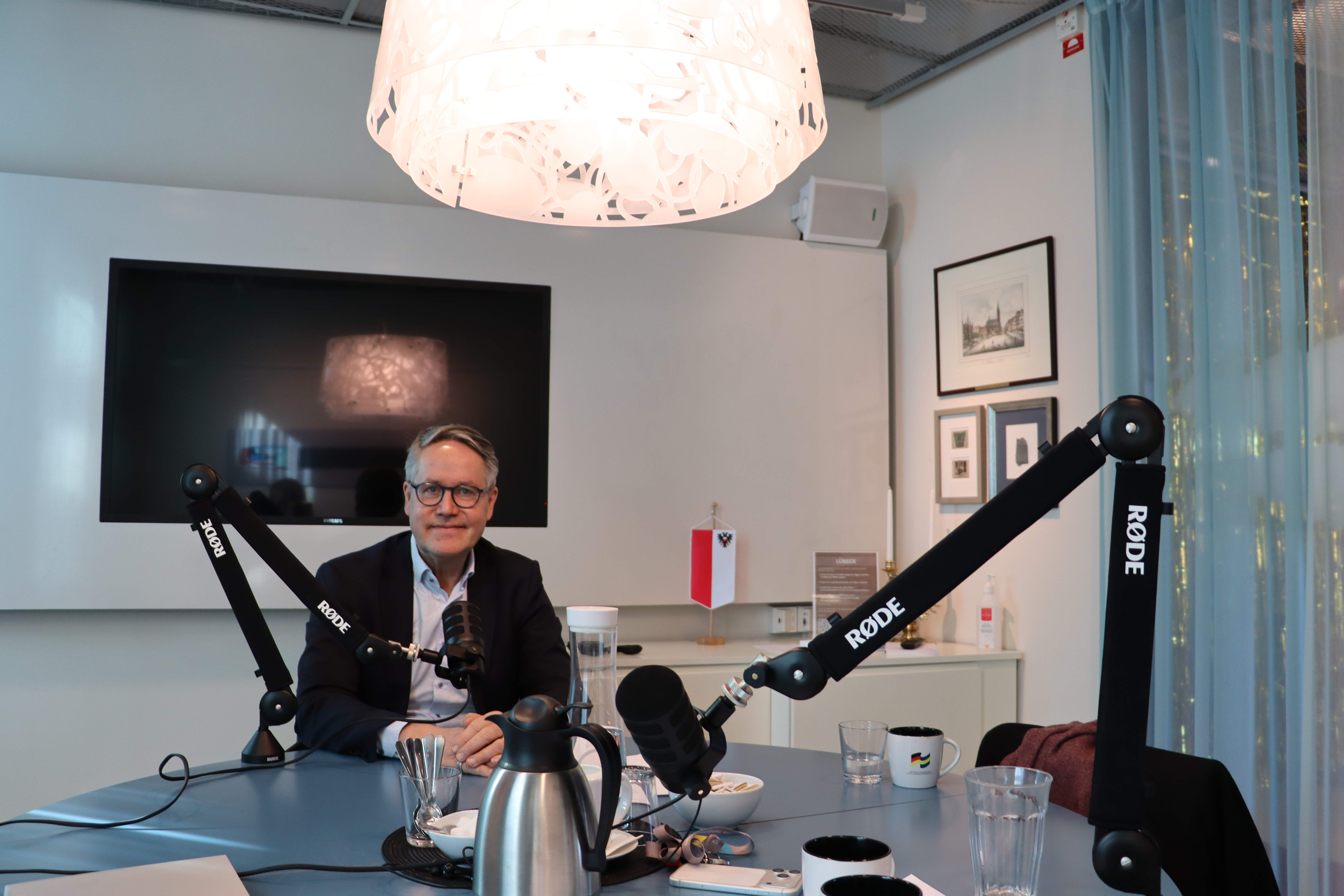 Ett mötesrum med ett runt bord där mikrofonarmar och inspelningsutrustning är uppställda. På bordet står glas, kaffemuggar och en termos. I bakgrunden syns en stor väggmonterad skärm, flera inramade bilder och ett fönster med gardiner. En person sitter vid bordet, och en taklampa hänger över inspelningsytan
