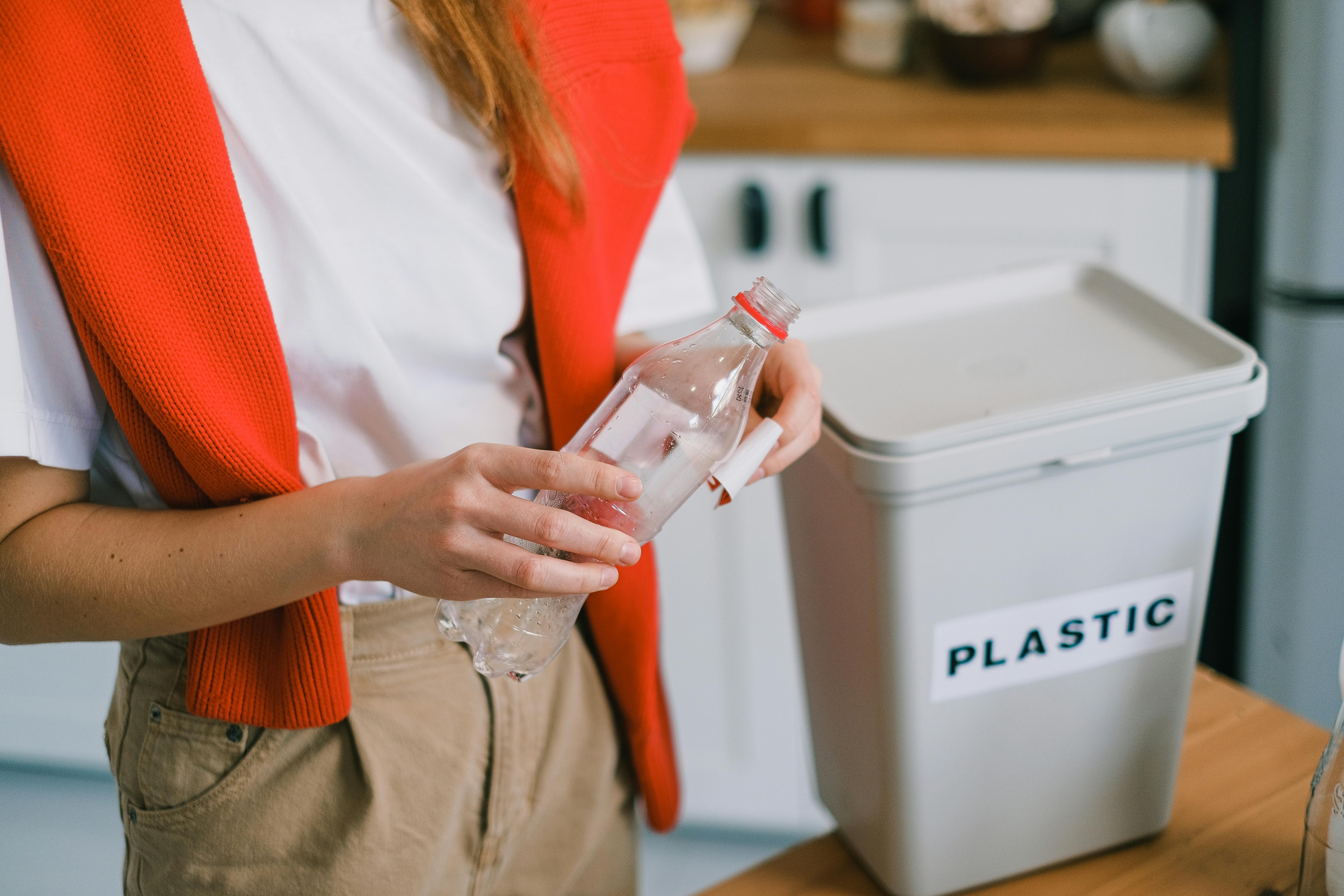 Person håller en tom plastflaska och står bredvid en behållare märkt ”PLASTIC”, redo att återvinna flaskan i ett kök.