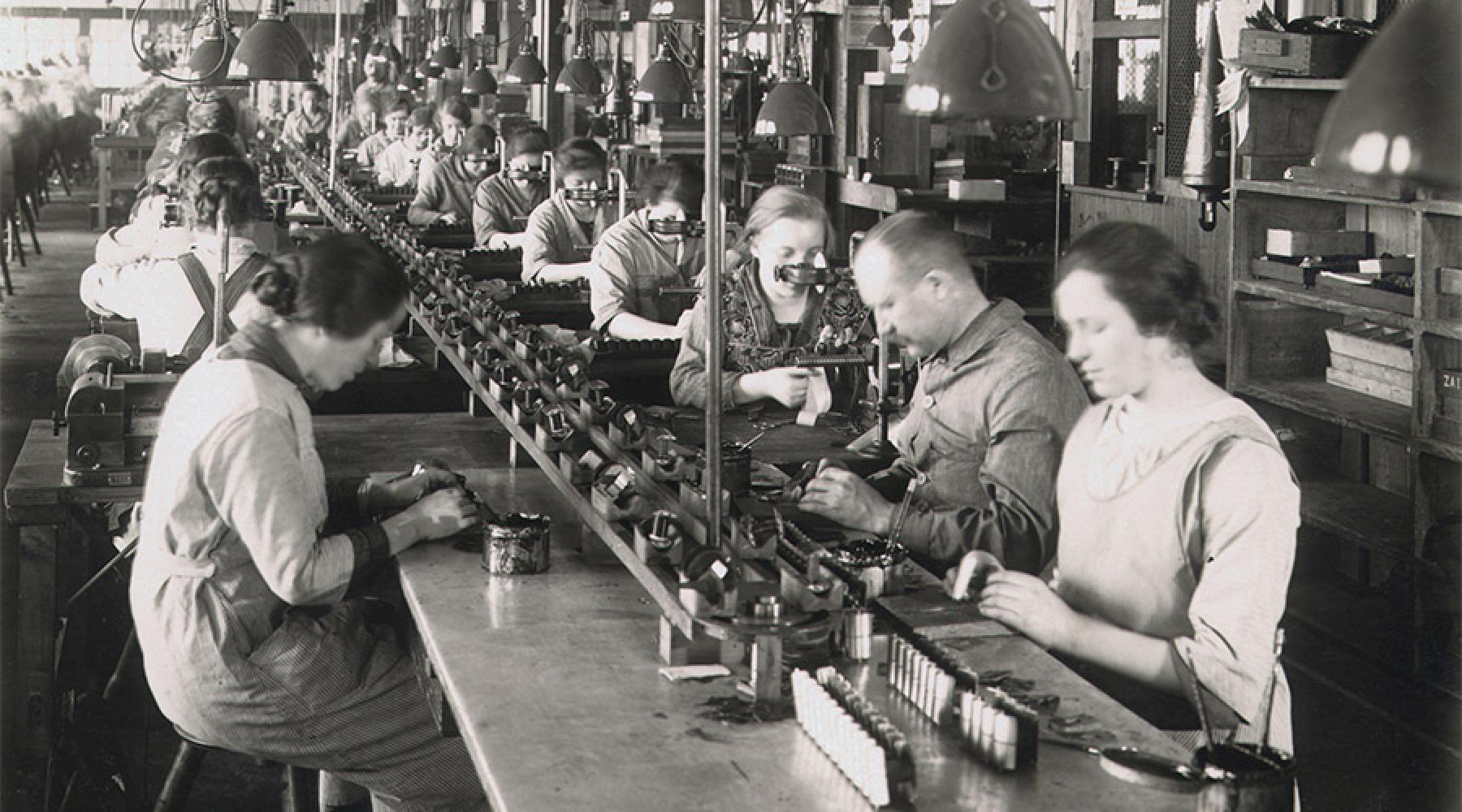 Historiskt svartvitt fotografi av en fabrik med en lång monteringslinje. Flera personer sitter eller står vid arbetsstationer och utför manuella arbetsuppgifter med komponenter och maskiner. Produktionshallen är utrustad med arbetslampor och industriell utrustning.