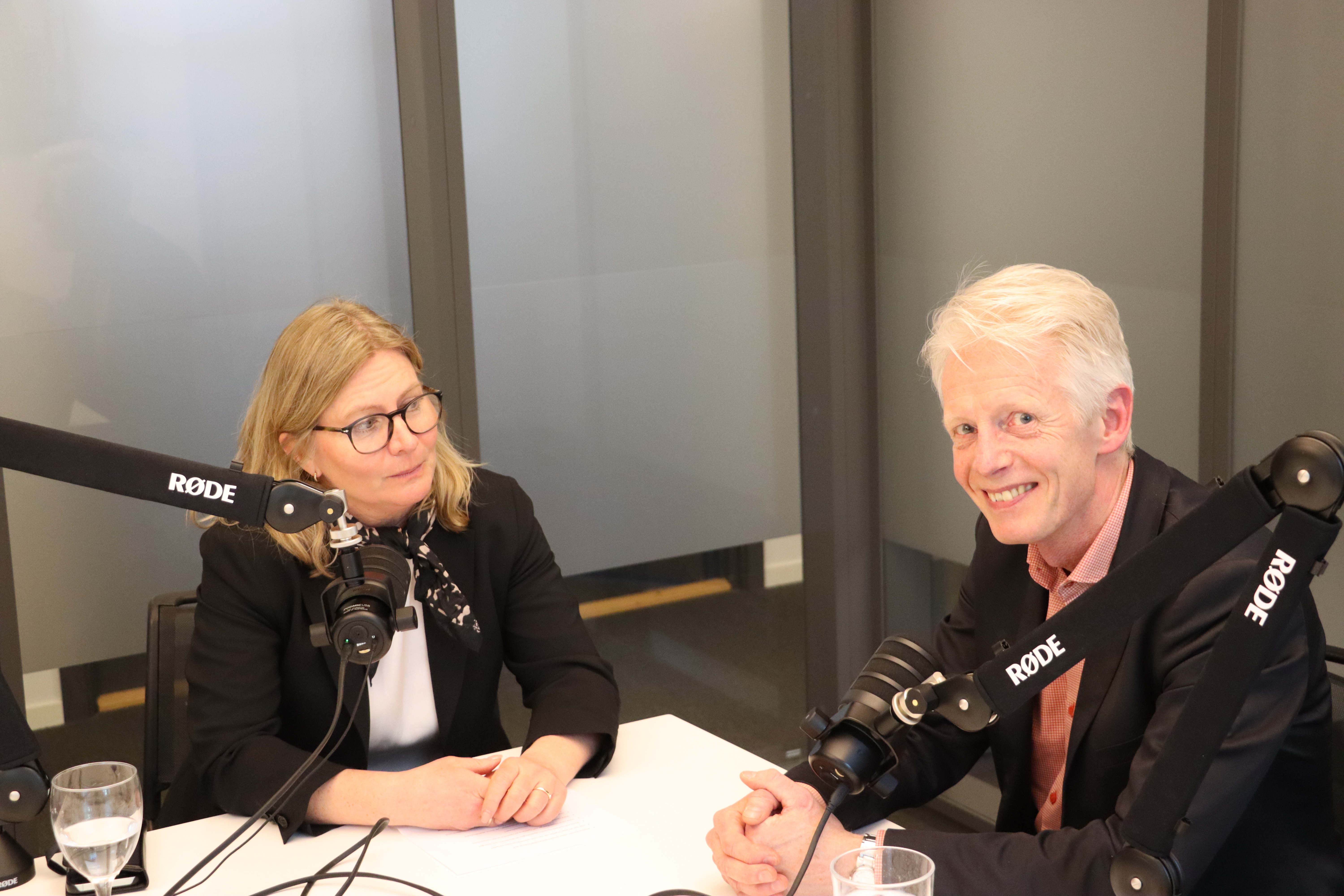 Två personer sitter vid ett bord i en studio och talar framför mikrofoner monterade på rörliga mikrofonarmar. På bordet står glas med vatten och ett block. Bakgrunden består av väggar med stora glaspartier, vilket ger rummet ett modernt och professionellt intryck. Det är en inspelningssituation för en podcast.