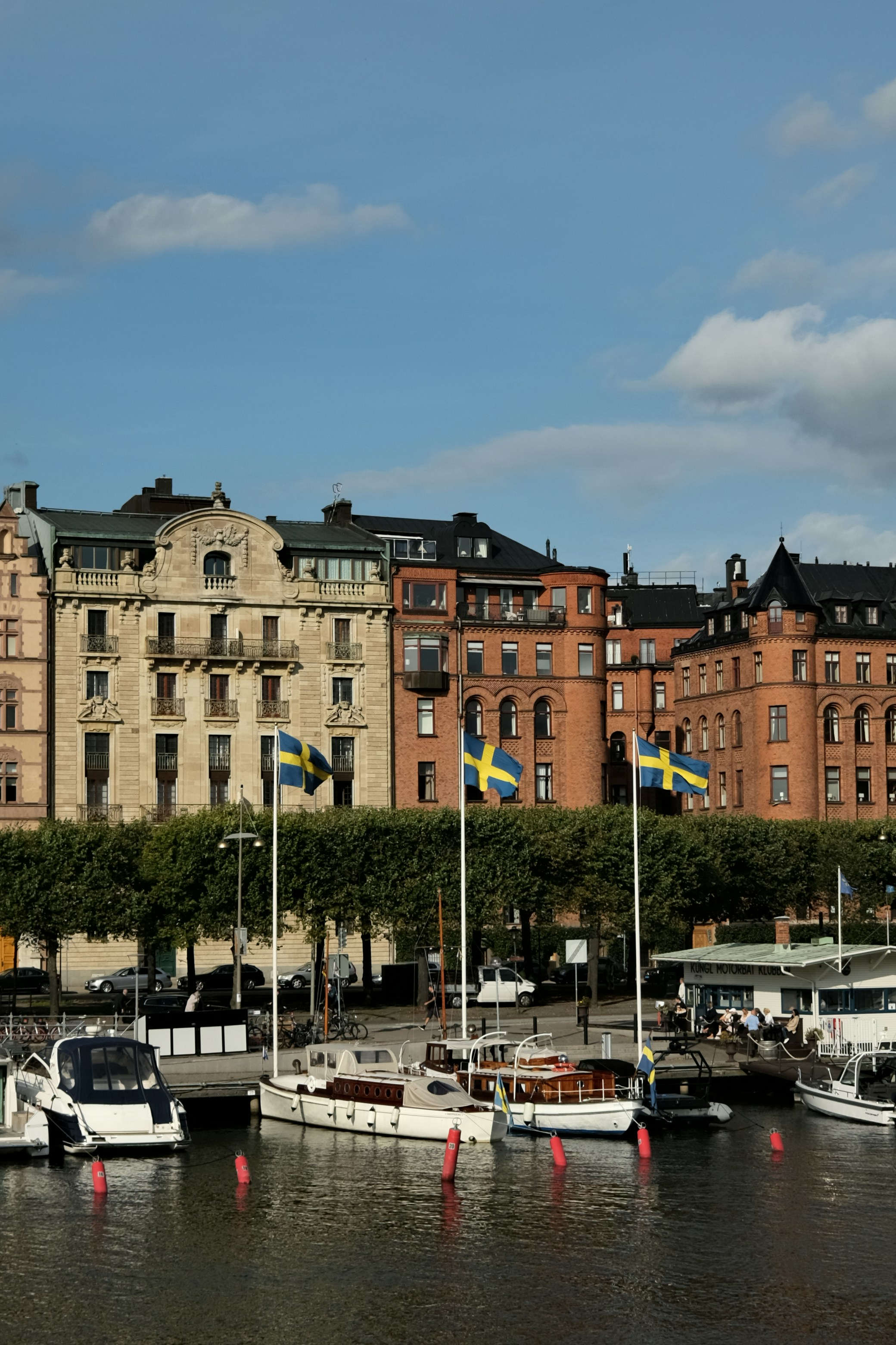 Boats moored along a waterfront with rows of historic multi‑story buildings and trees in the background under a partly cloudy sky Sweden 