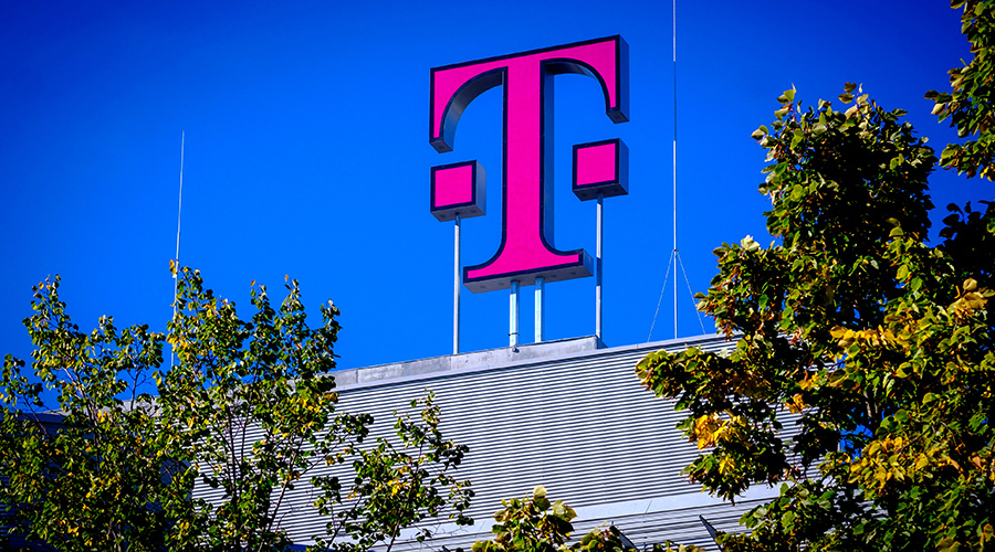 Ein großes magentafarbenes T‑Logo auf dem Dach eines Gebäudes, umgeben von Bäumen und klarem blauem Himmel.