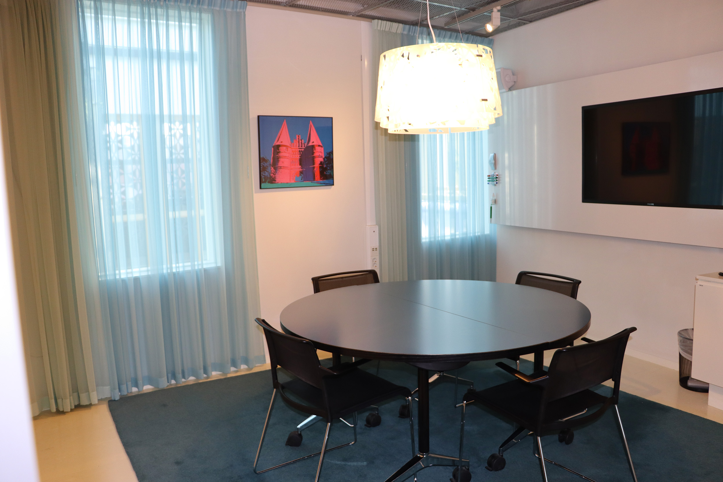 Heller Konferenzraum in der Deutsch-Schwedischen Handelskammer mit rundem Tisch, vier Stühlen, wandmontiertem Fernseher und großen Fenstern mit hellen Vorhängen.