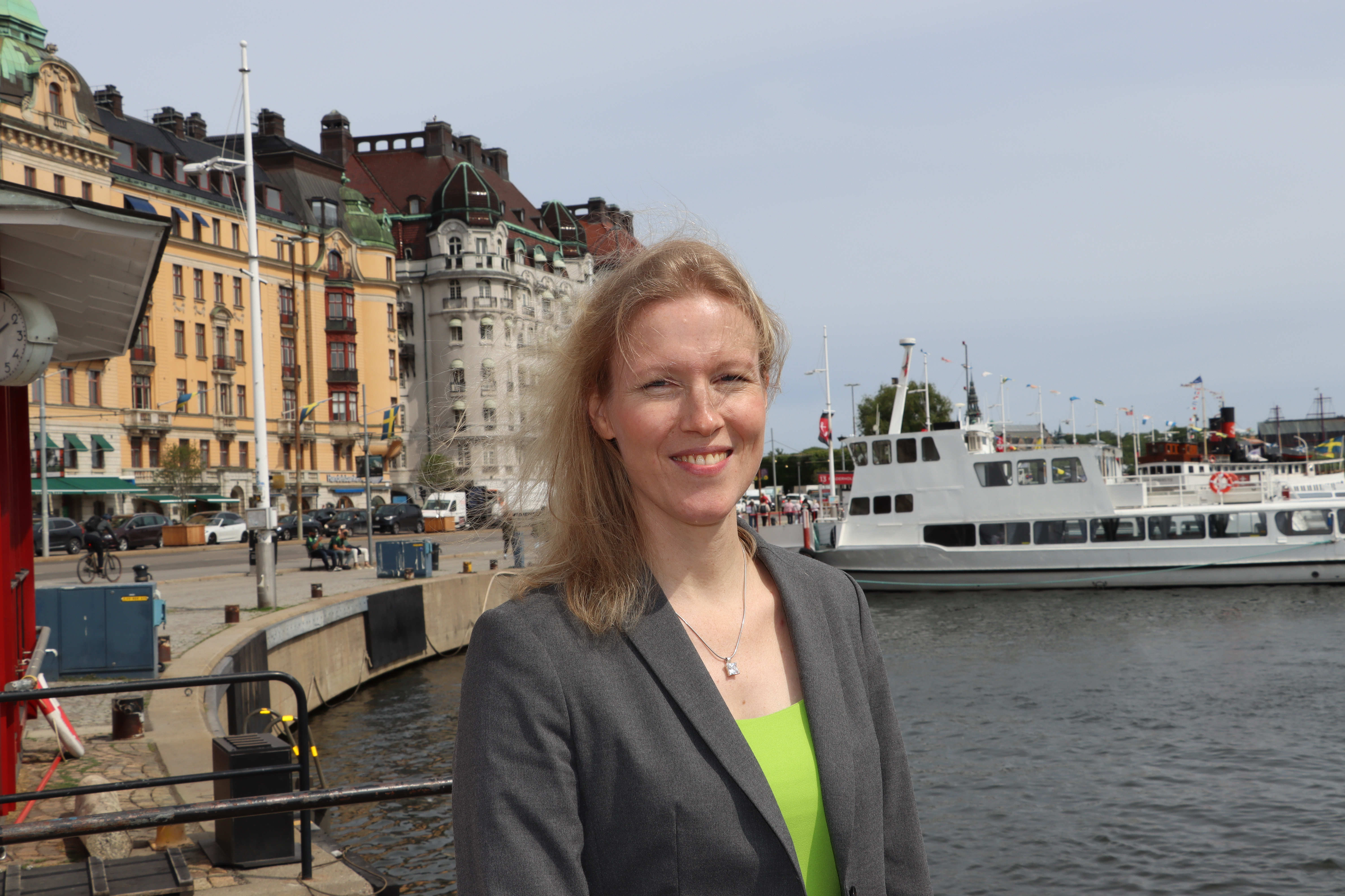 En person står vid en kaj i en stadsmiljö med vattnet och flera vita passagerarbåtar i bakgrunden. Längs kajen syns färgglada byggnader i klassisk arkitektur, och himlen är lätt molnig.