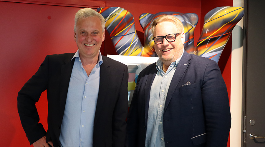 Zwei Personen in Blazern stehen vor einer roten Wand mit einem farbenfrohen, abstrakten Kunstwerk im Hintergrund.