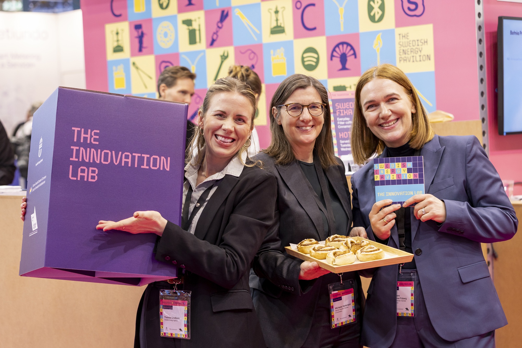 På plats på mässan E-world energy & water i den svenska energipaviljongen, tre medarbetare håller upp en kub med monterns budskap pch en bricka med kanelbullar.