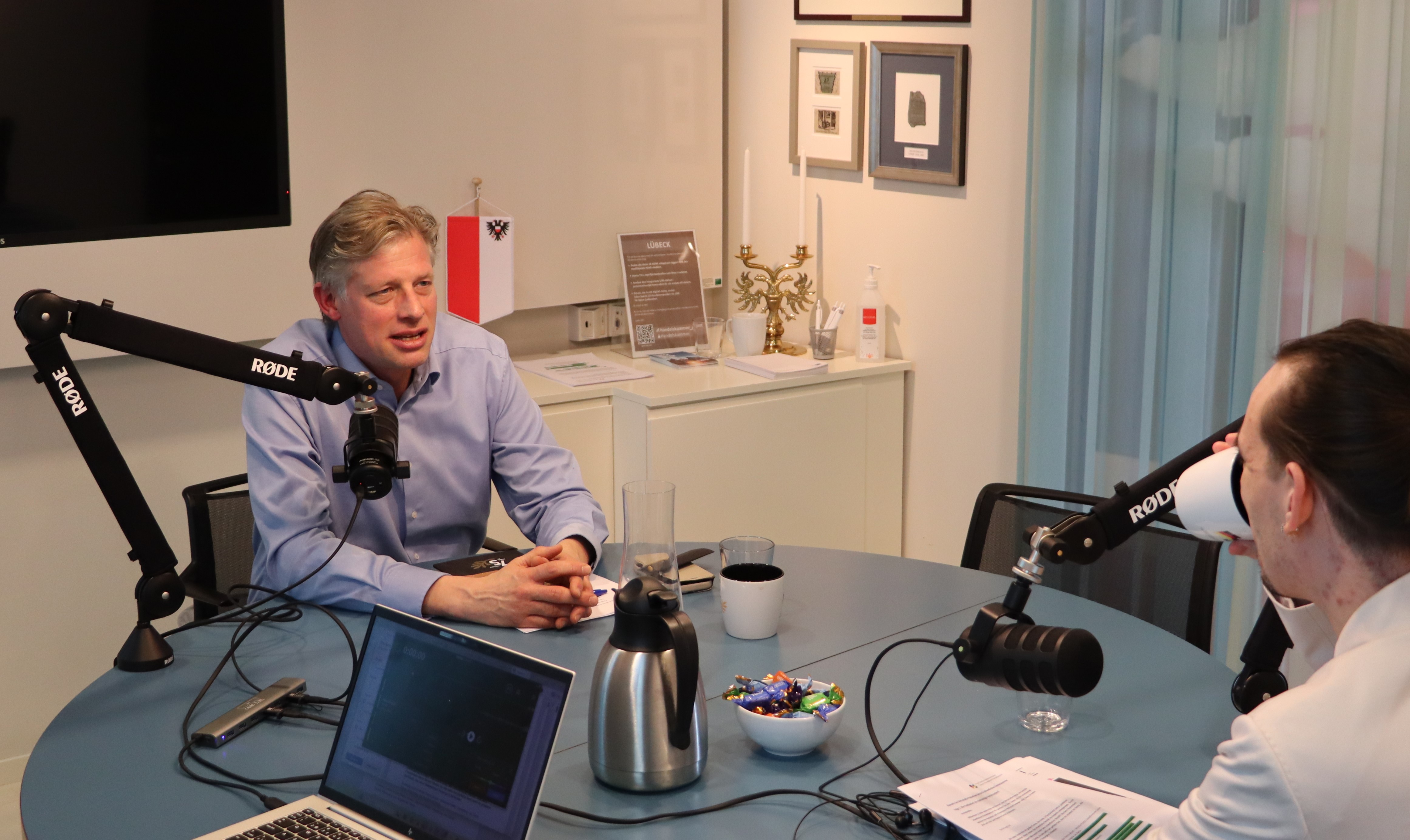 Två personer sitter vid ett runt bord i en studio och spelar in ett samtal med mikrofoner monterade på svängbara armar. På bordet finns en kaffekanna, en kopp, papper, ett glas och en bärbar dator. I bakgrunden syns en hylla med dekorationer och en vägg med inramade bilder.