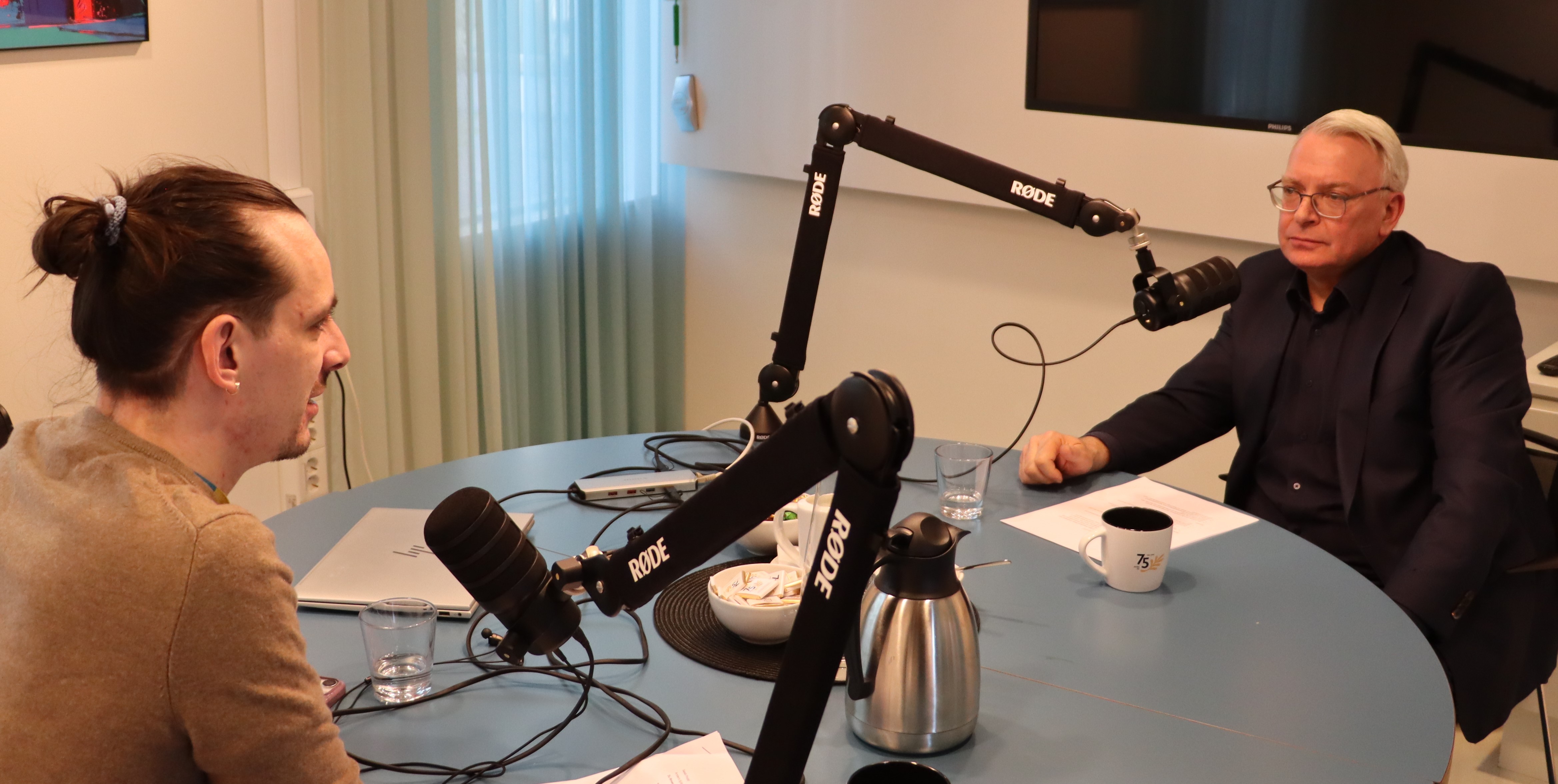 Två personer sitter vid ett runt bord i en studio och spelar in ett samtal med mikrofoner monterade på svängbara armar. På bordet finns en kaffekanna, en kopp, glas, papper och en bärbar dator. I bakgrunden syns en TV‑skärm och ett fönster med ljusinsläpp.