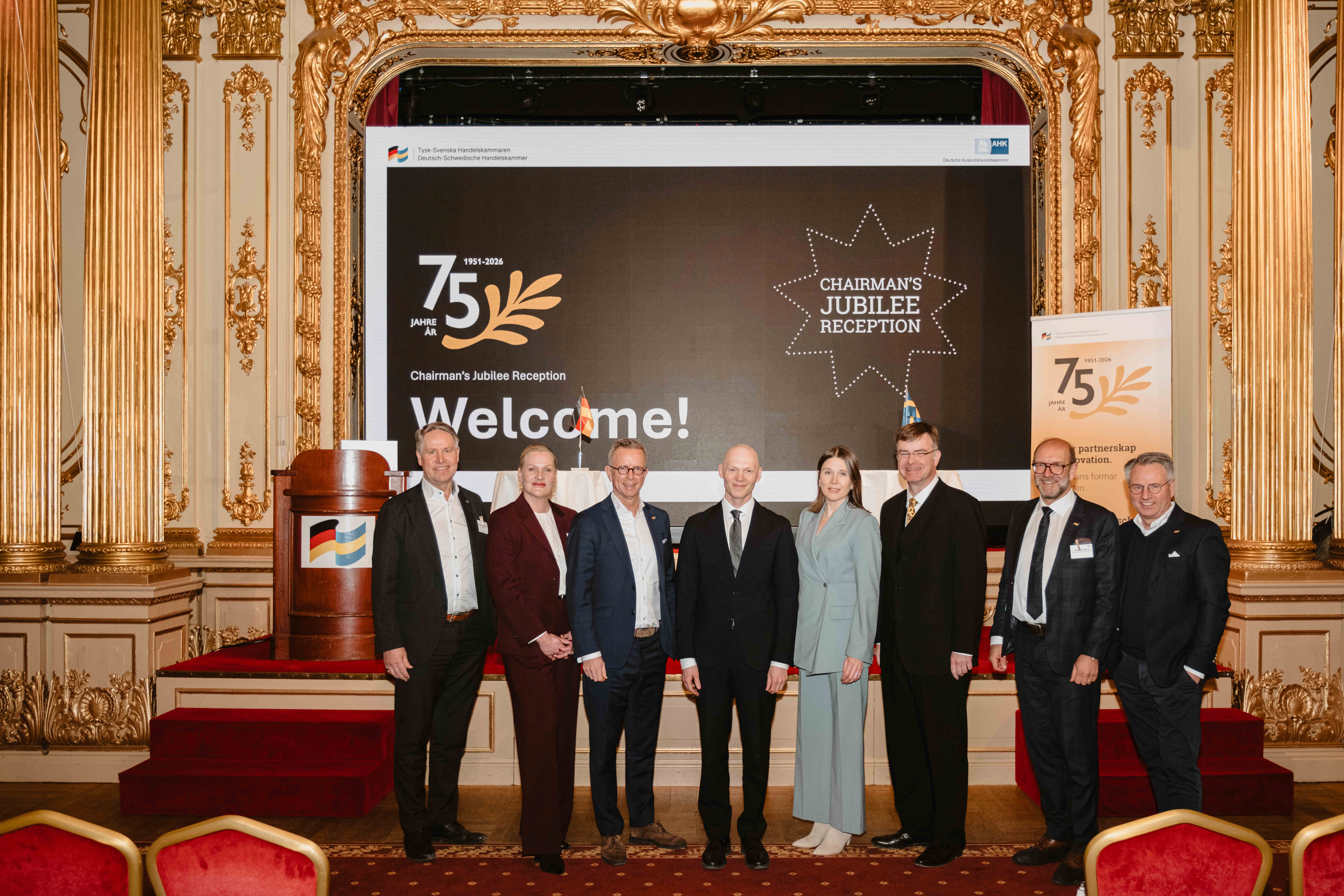 Gruppfoto på scen under ett 75‑årsjubileumsevent med dekorativ salongsmiljö, stor presentationsskärm och välkomstbudskap i bakgrunden