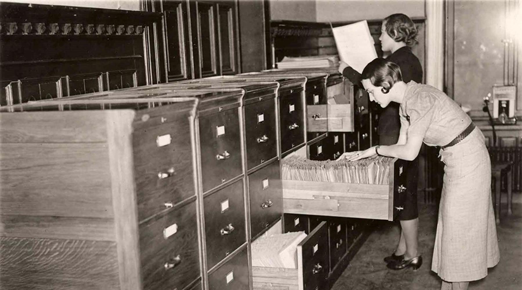 Historiskt svartvitt fotografi av ett arkiv med flera kortkatalogskåp i trä. Två personer söker igenom öppna lådor med dokument och registerkort i ett rum med hyllor i bakgrunden.