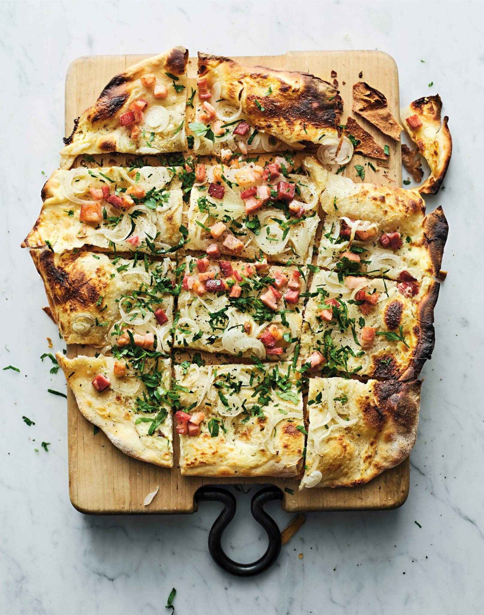  En rektangulär flammkuchen ligger på en träbräda med handtag på ett ljust bord. Flammkuchenen är skuren i rutor och toppad med skivor av potatis, smält ost, små tärningar av kött samt färska gröna örter. Kanterna är tunna, gyllenbruna och lätt brända på vissa ställen. Ge feedback om BizChat