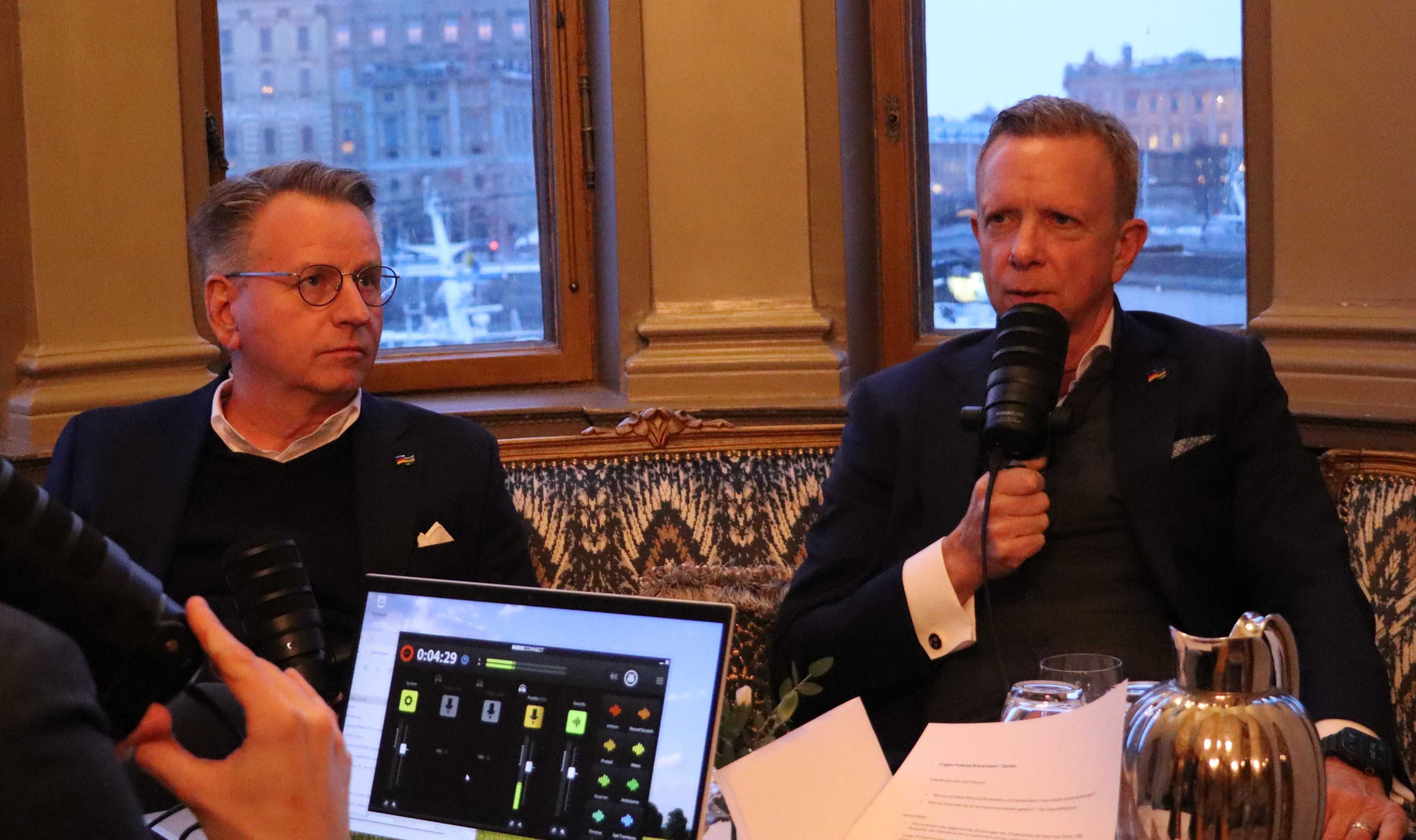 Två personer sitter vid ett bord i en elegant inomhusmiljö och håller mikrofoner under en inspelning. Framför dem finns ett bord med dokument, ett glas, en kanna och en bärbar dator med inspelningsprogram öppet. Ett stort fönster i bakgrunden visar en stadsmiljö vid vatten.