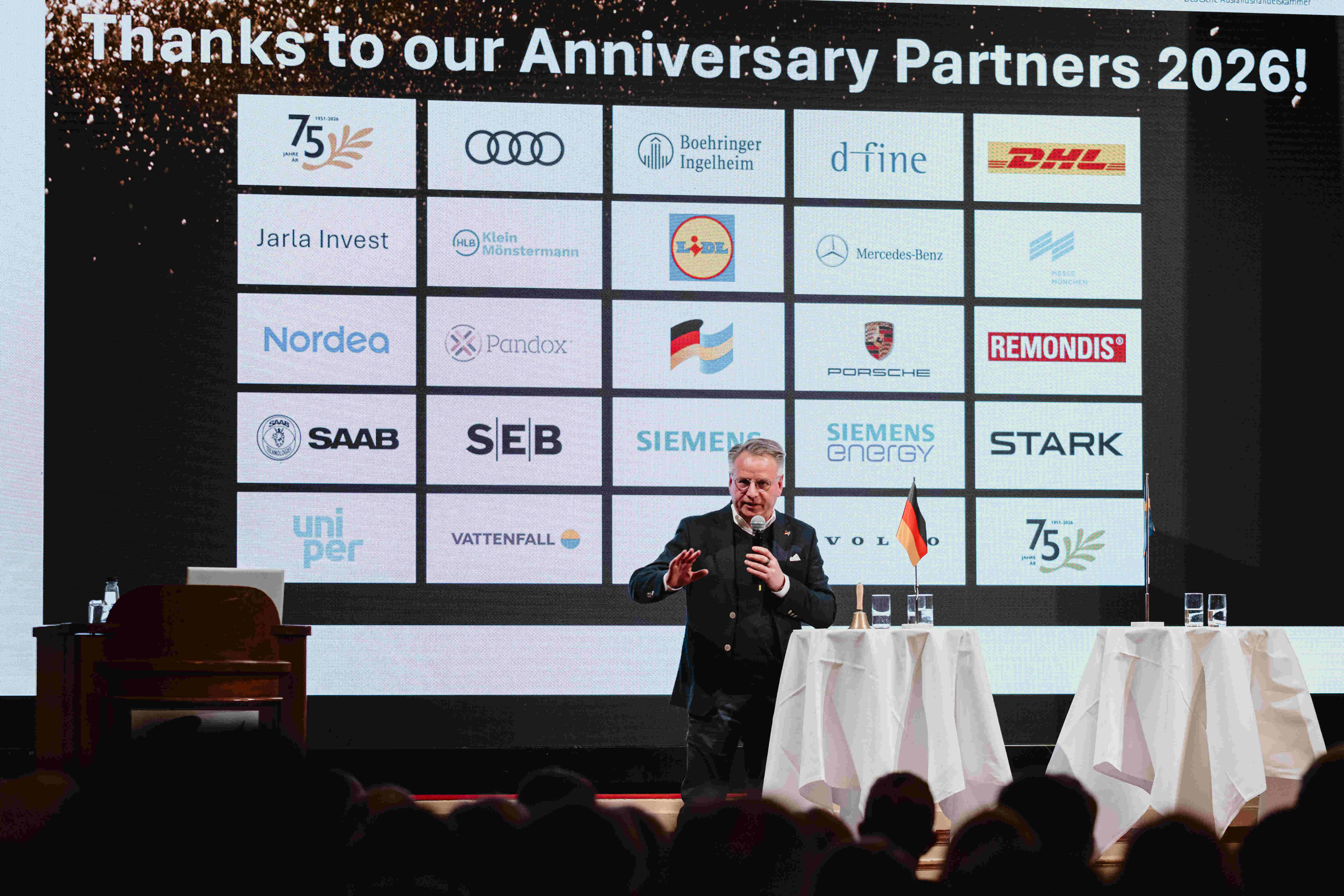 Talare på scen under ett jubileumsevent med logovägg som visar partnerföretag, inkluderande stora internationella varumärken, samt tysk och svensk flagga i förgrunden
