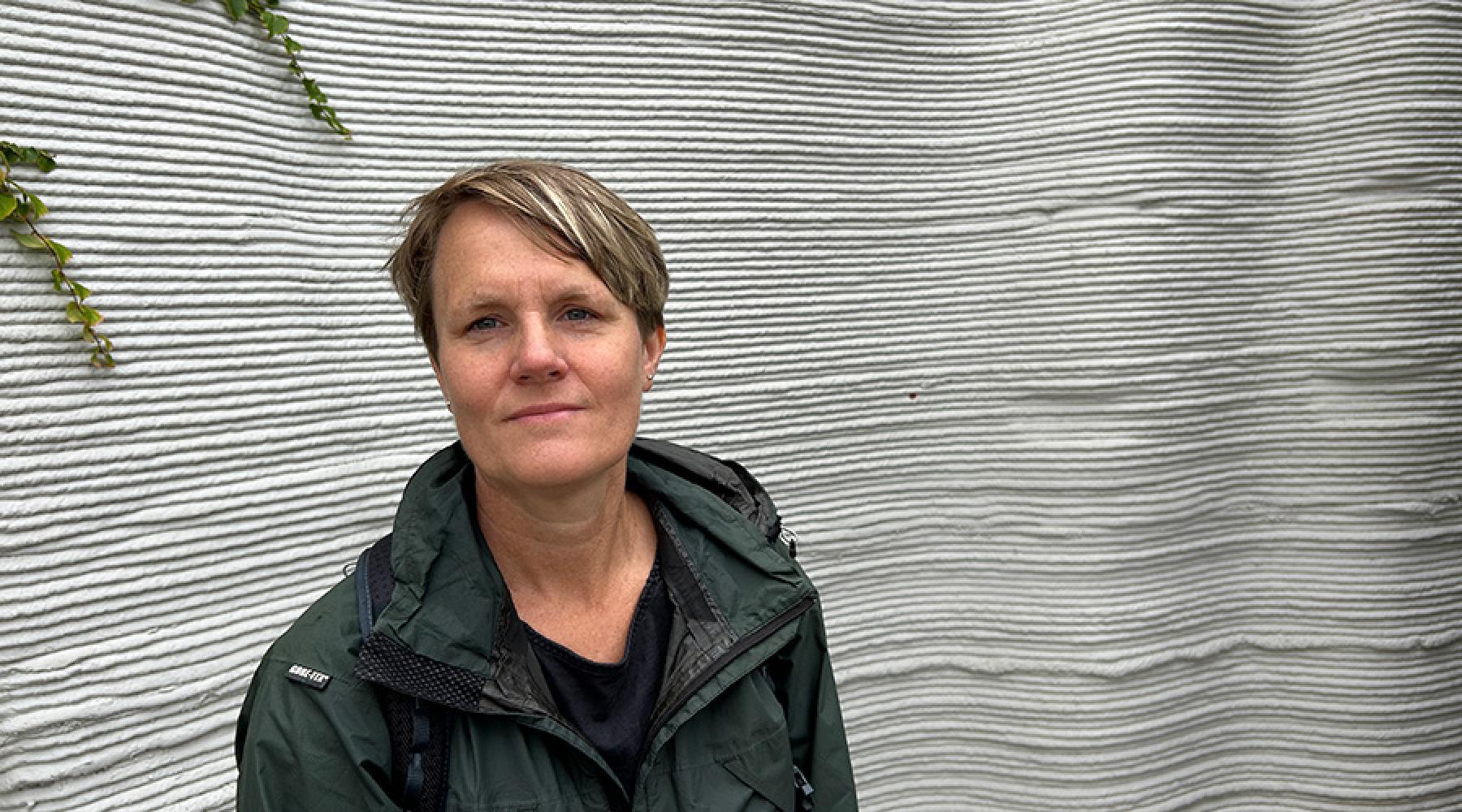 En person står framför en vägg med horisontella vågformade strukturer och bär en mörk jacka.