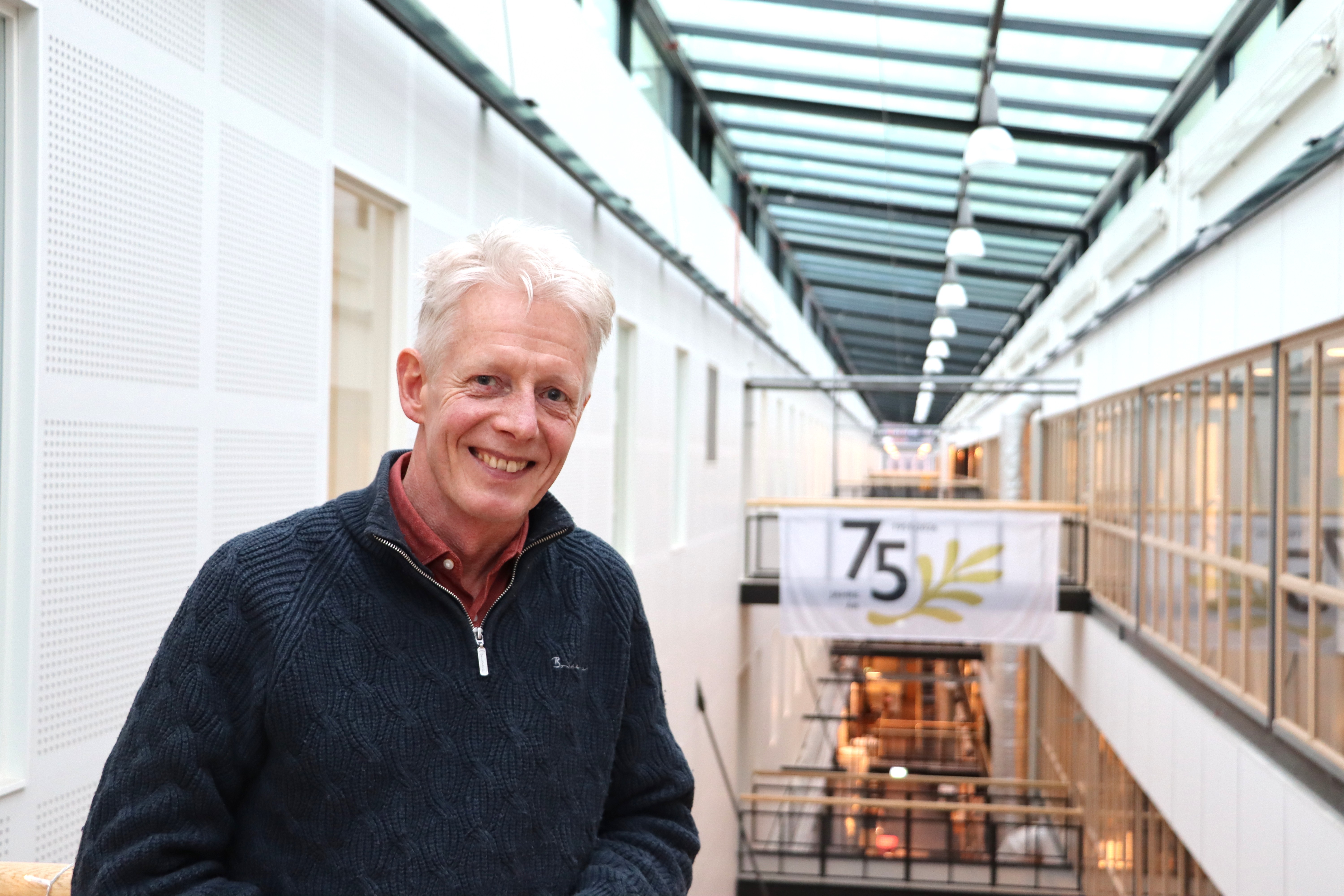 En person står inomhus i en ljus, lång korridor med glasat tak. I bakgrunden hänger en stor vit banner med texten “75” och en gul dekorativ gren