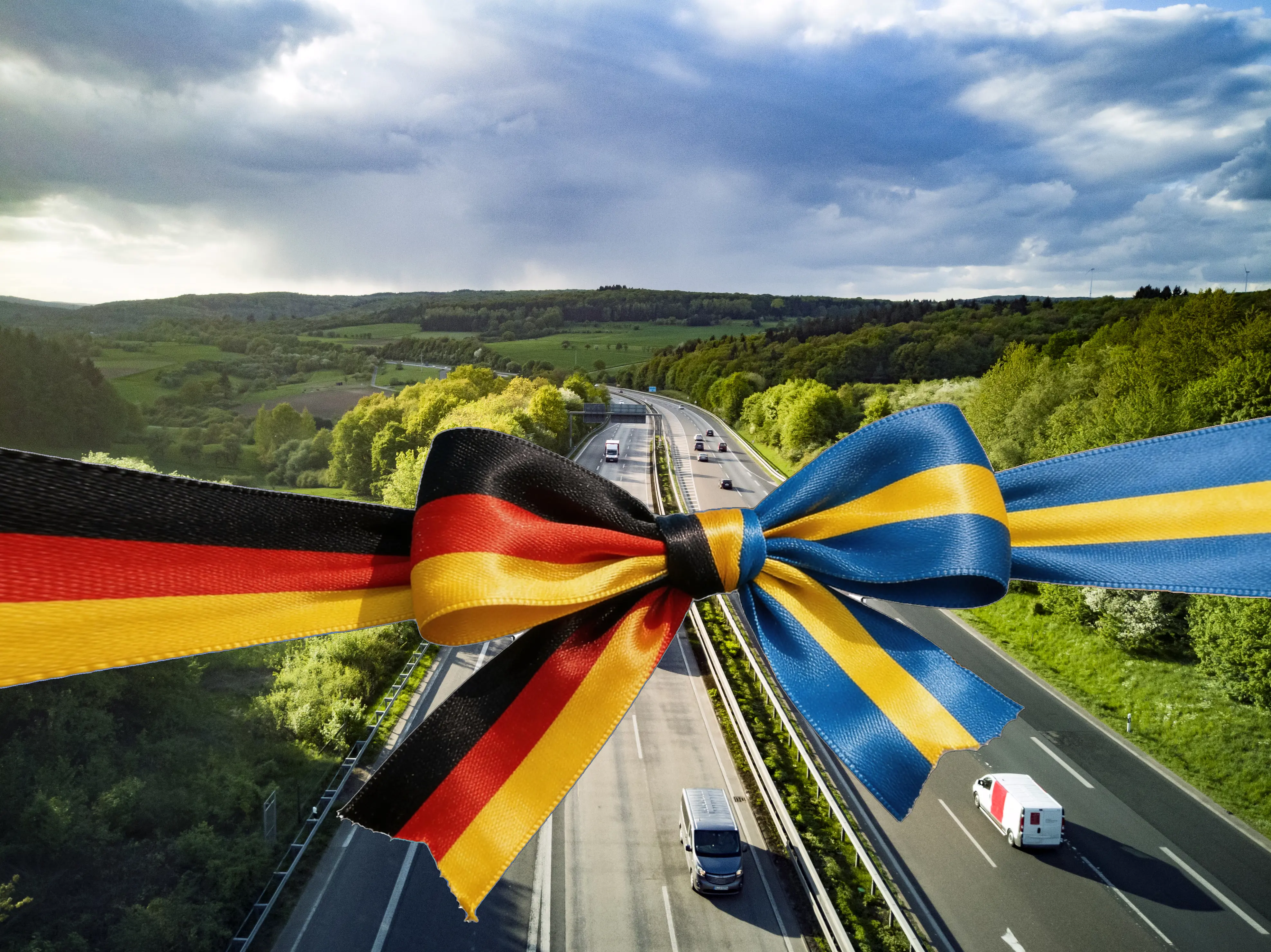 Ett landskap med en motorväg som löper genom gröna skogar och kullar, med en stor digitalt placerad rosett i tyska och ukrainska flaggans färger över vägen.