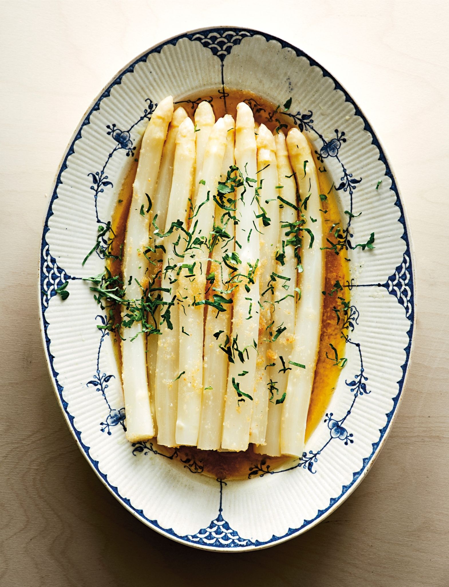 Vita sparrisar ligger tätt upplagda på ett ovalt fat med blå dekor. Sparrisarna är täckta med en gyllene sås och garnerade med finhackade gröna örter. Fatet står på en ljus bordsyta och ljuset är mjukt, vilket framhäver sparrisens glansiga yta och såsens färg.