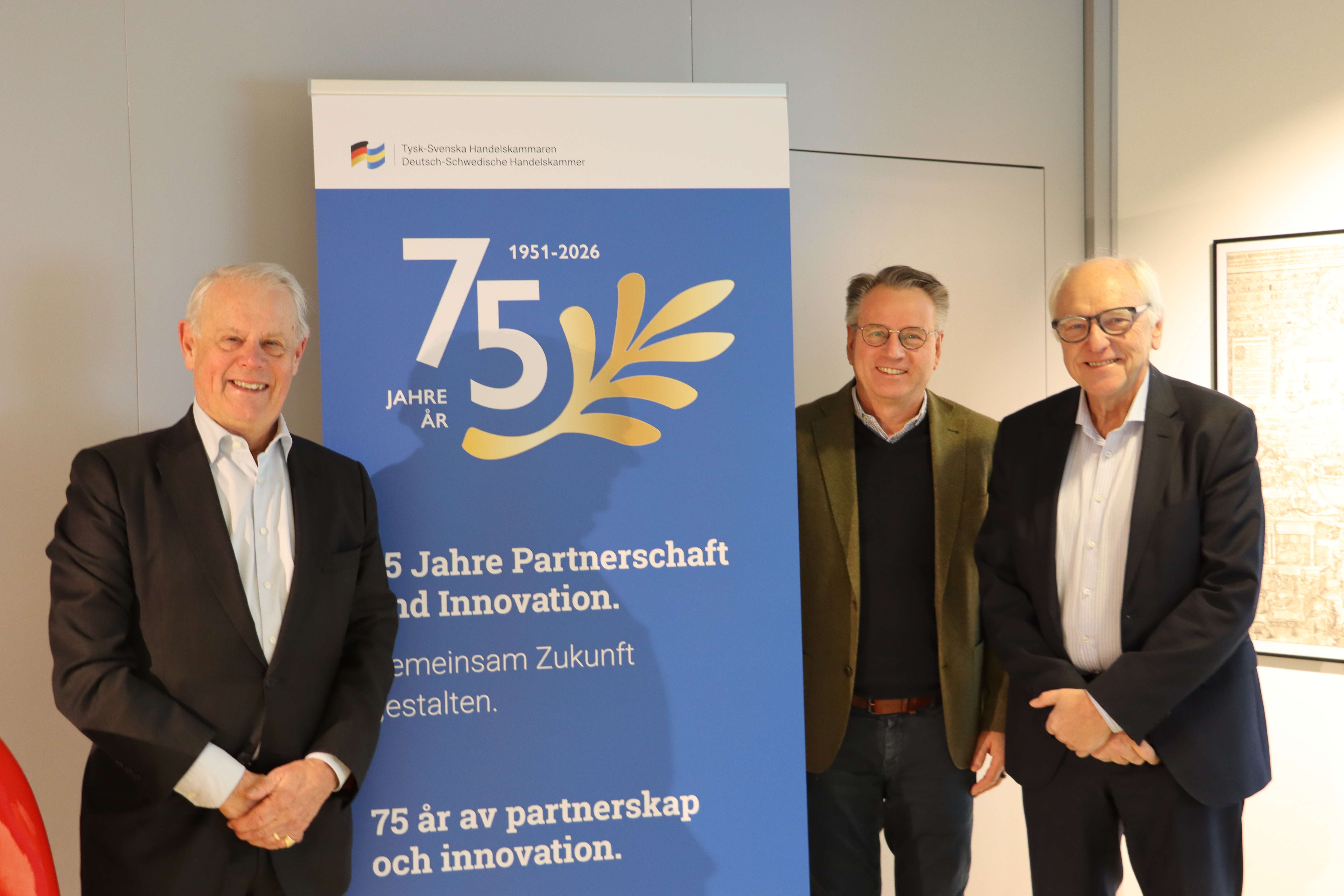 Tre personer står bredvid ett blått jubileumsställ med texten ’75 år av partnerskap och innovation’ i en inomhusmiljö.