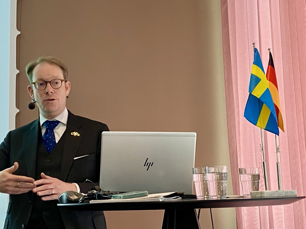 En person står vid ett högt bord med en öppen laptop framför sig och håller en presentation. Bredvid datorn står tre glas vatten och två flaggor – en svensk flagga och en tysk flagga – placerade i ett stativ. Bakgrunden består av en ljus vägg och ett rosa draperi