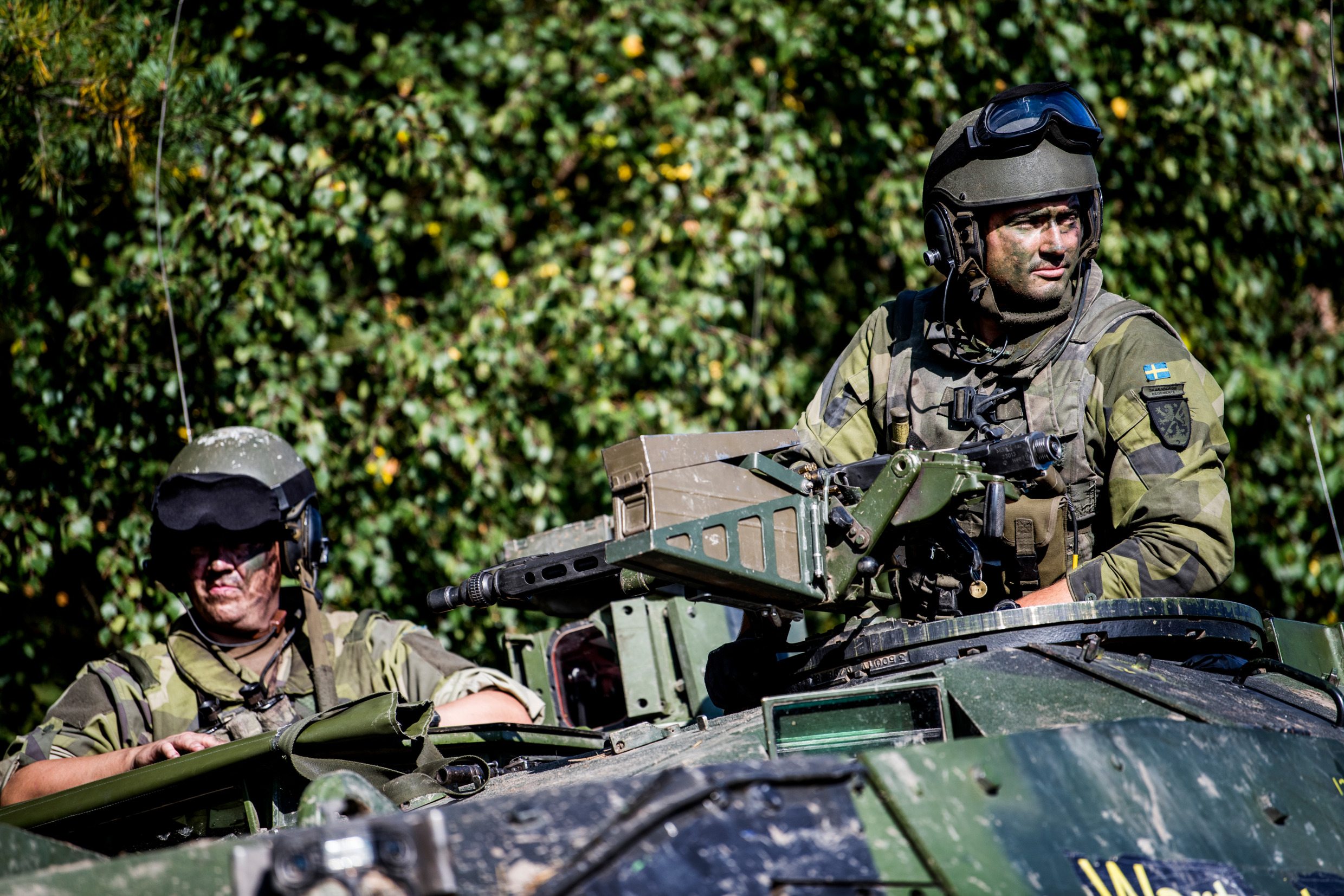 Två uniformerade personer sitter på ett militärfordon omgivet av grönska, med en monterad kulspruta och övrig utrustning synlig på fordonets ovansida