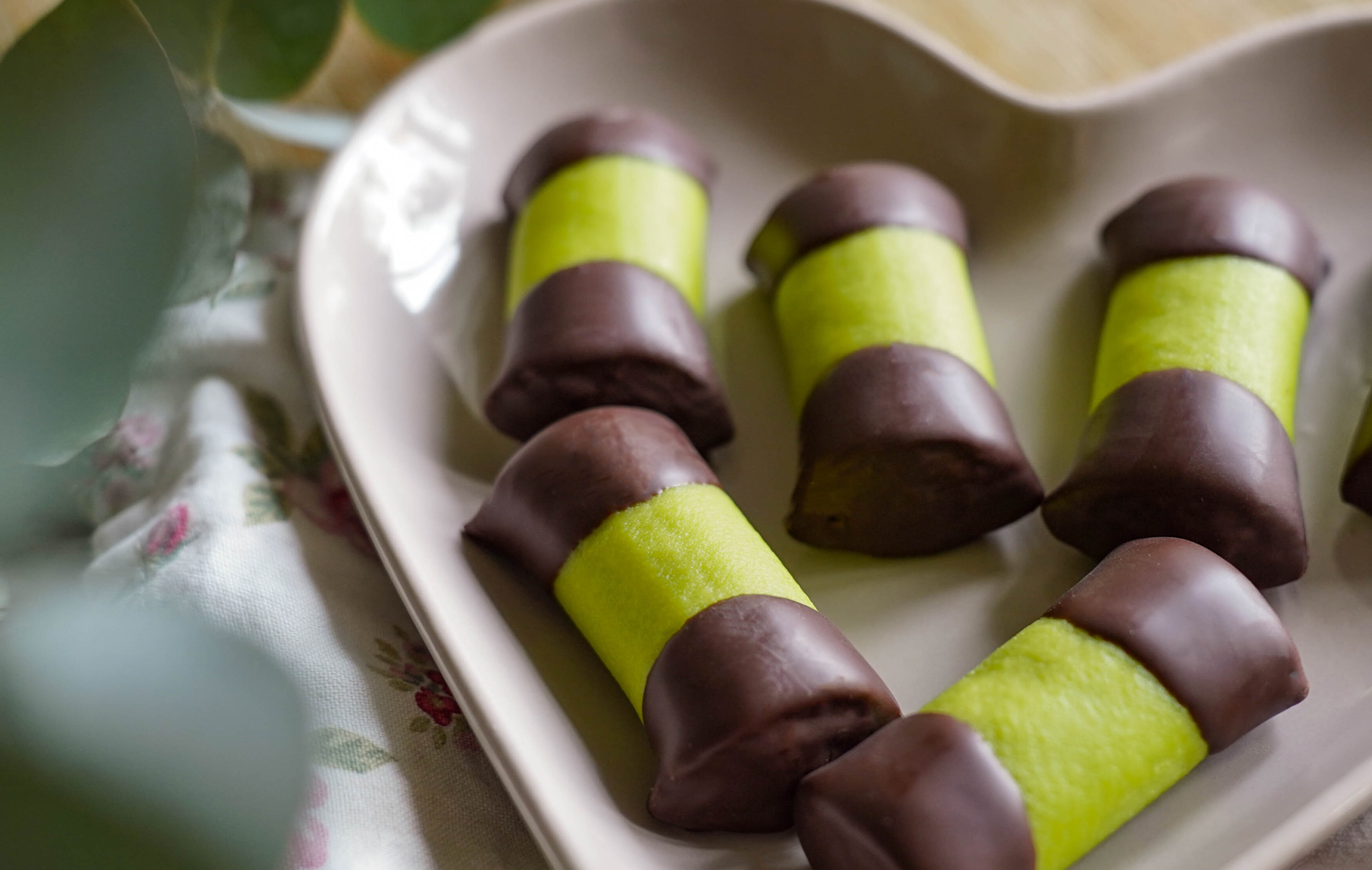 Mehrere Punschröllchen mit grünem Marzipan und Schokoladenüberzug liegen auf einer Servierplatte auf einem gedeckten Tisch. Ge feedback om BizChat