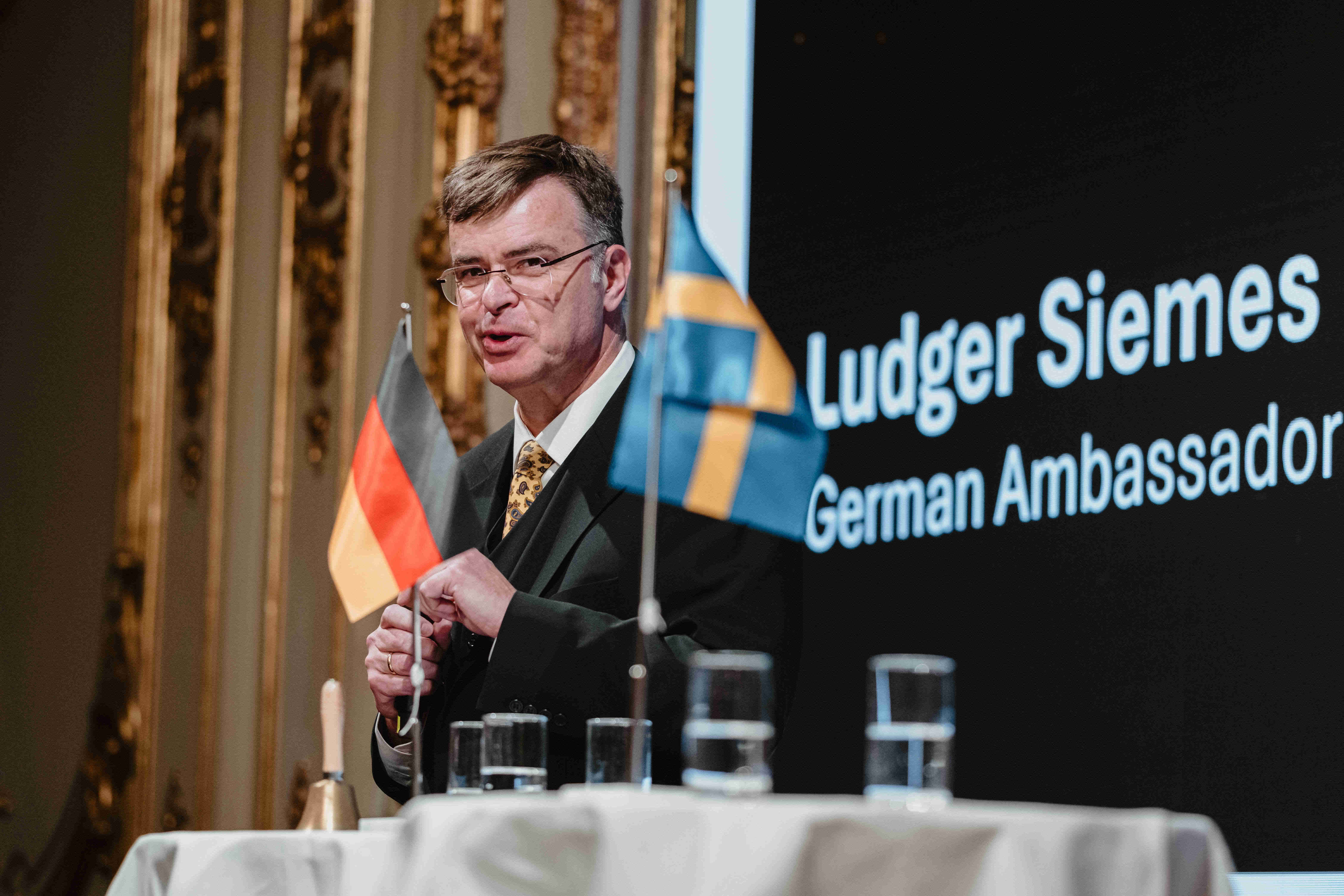 Talare står vid ett podium under ett tyskt‑svenskt evenemang, med tyska och svenska flaggor och en stor presentationsskärm i bakgrunden