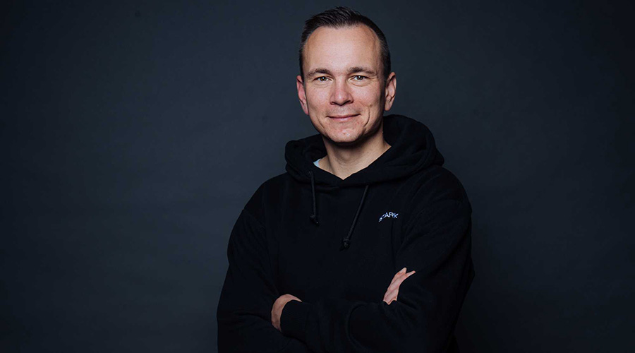 A person wearing a dark hooded sweatshirt stands against a dark background with arms crossed.