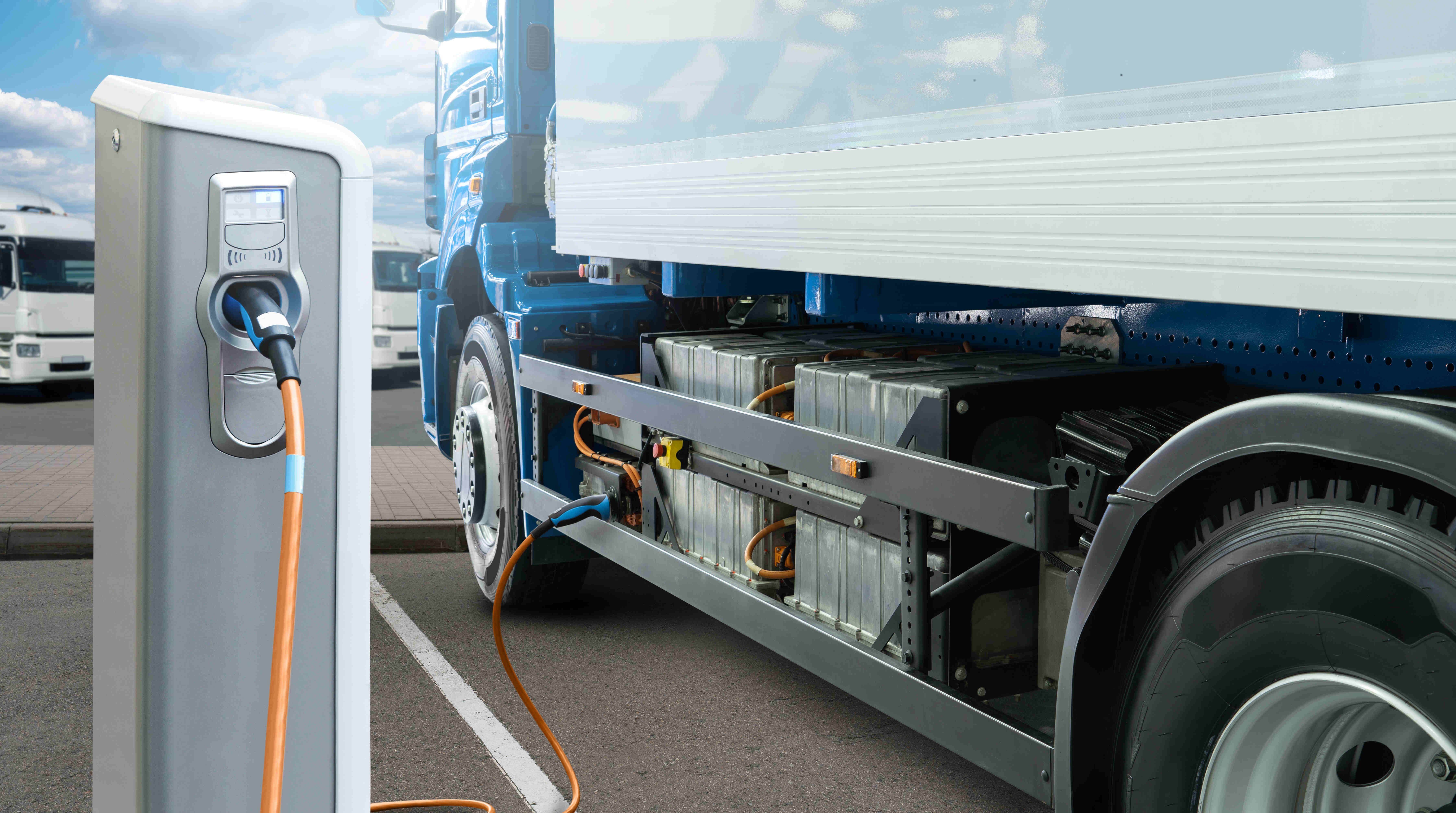 En eldriven lastbil står parkerad vid en laddstation utomhus. En laddkabel är ansluten till lastbilens batterisystem som är synligt längs fordonets sida, med andra lastbilar i bakgrunden på en logistikparkering.