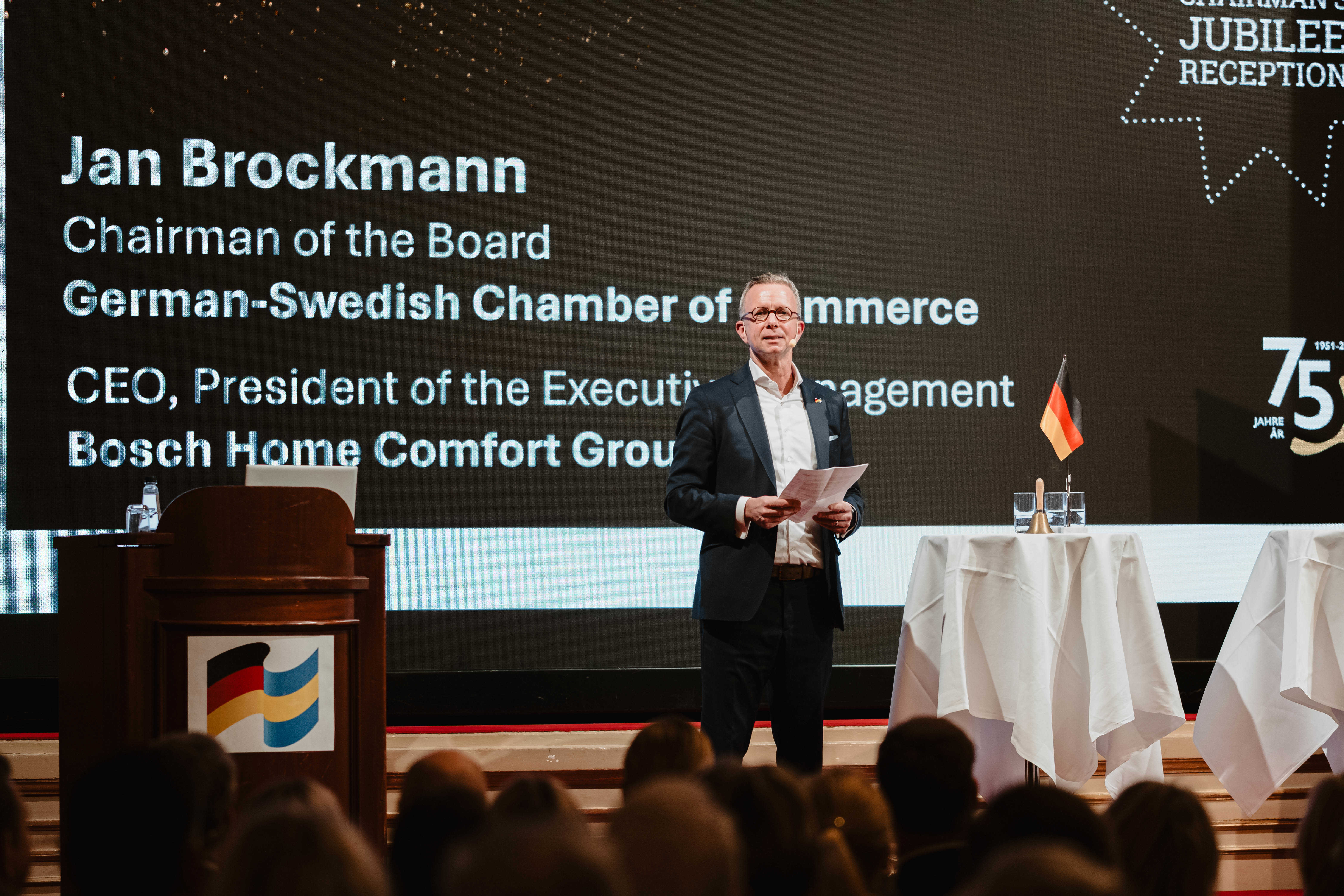 Talare på scen under ett jubileumsevent hos German‑Swedish Chamber of Commerce, med presentationsskärm och flaggdekorationer i bakgrunden.