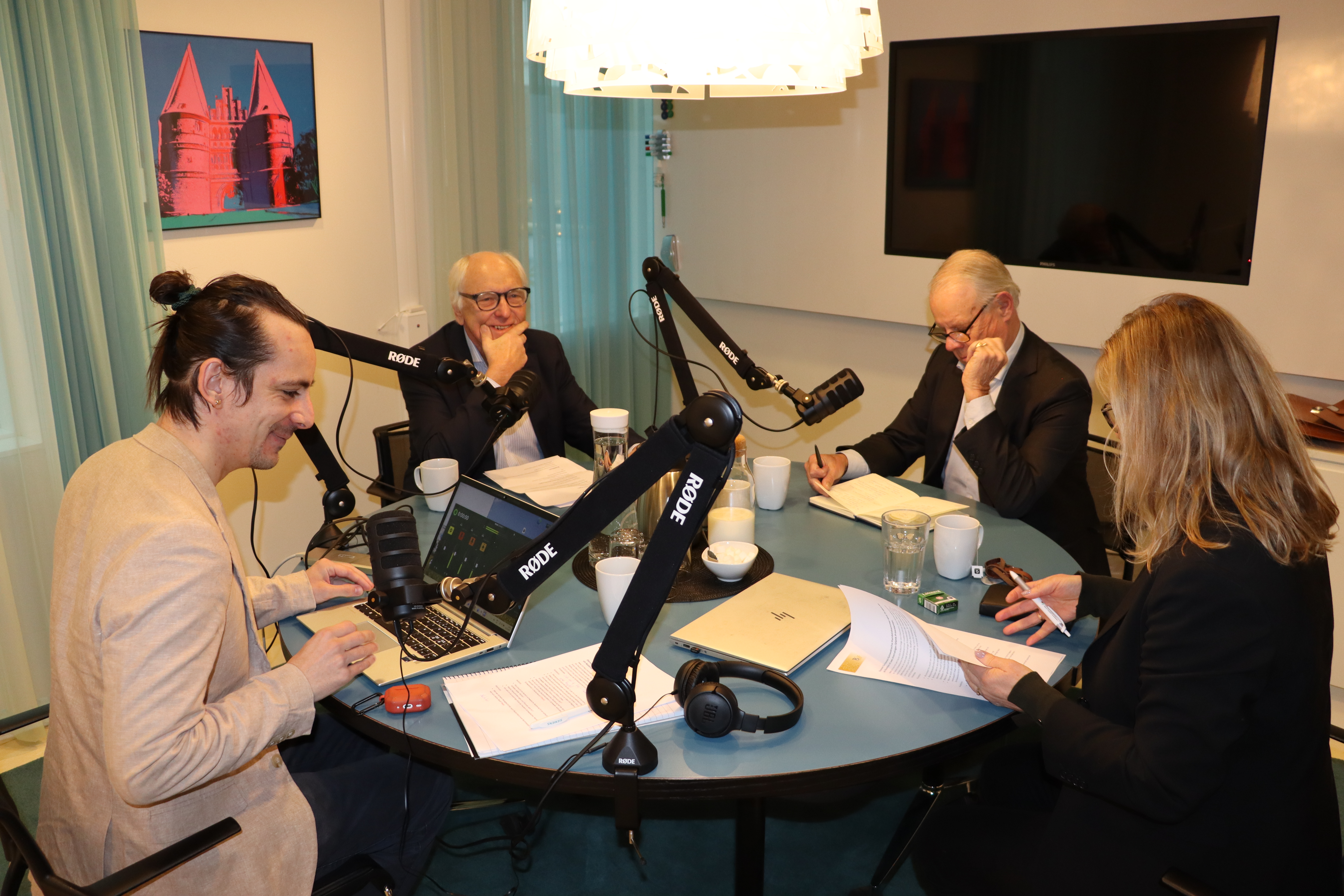 Fyra personer sitter runt ett bord med mikrofoner, dokument, en laptop och kaffekoppar i ett modernt mötesrum