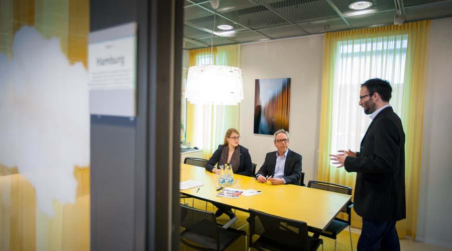 Två personer sitter vid ett bord och tittar på en talare som står vid bordets kortsida i ett av Tysk-Svenska Handelskammarens konferensrum.