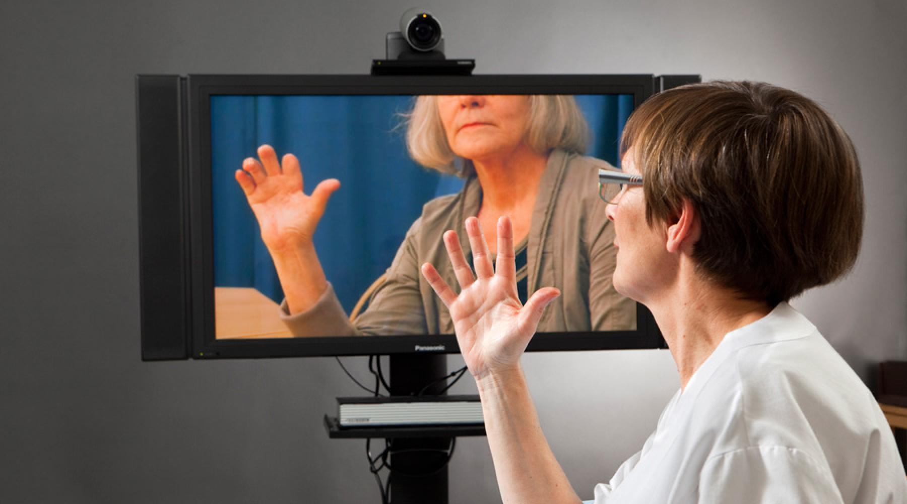 En läkare sitter framför en tv-skärm och håller upp en hand. På tv:n syns en annan kvinna som också håller upp handen i rehabiliteringssyfte.