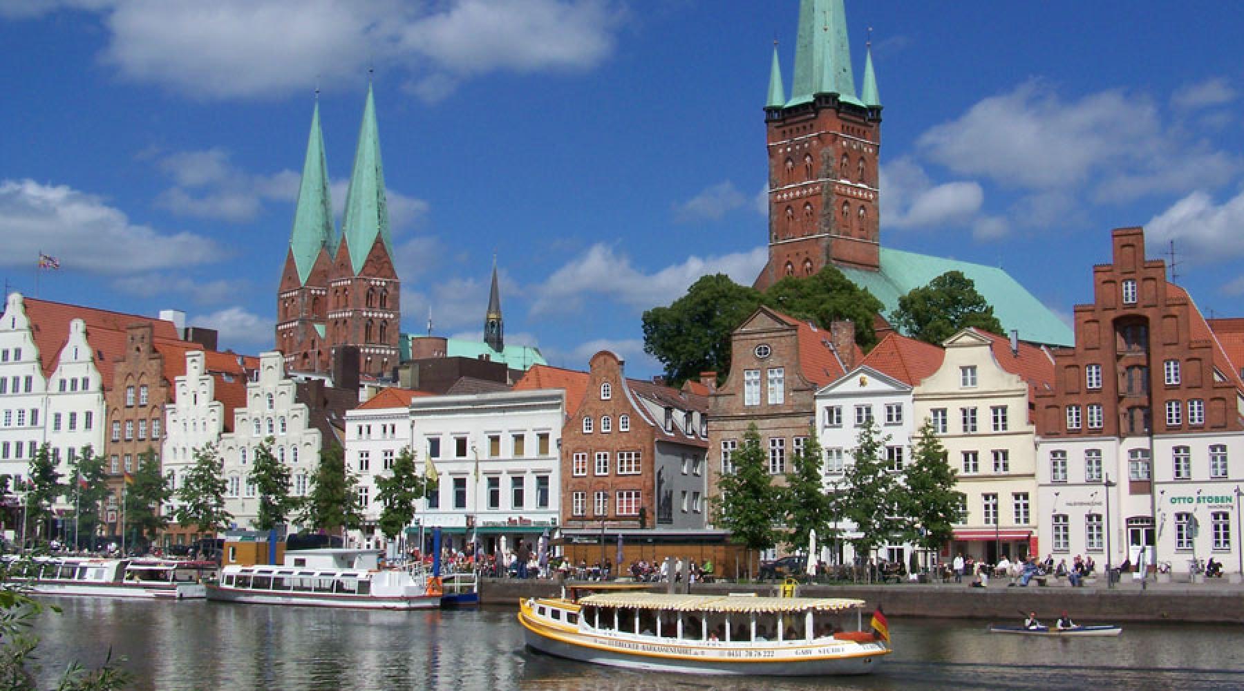 En gata med typiska tegel- och stenhus vid en kanal i Lübeck. På kanalen syns flera båtar, i bakgrunden syns två kyrkors torn.