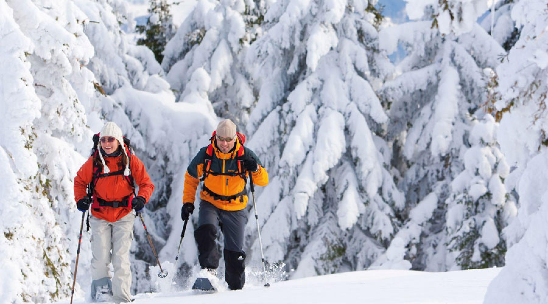 En kvinna och en man går på snöskor genom en skog som är täckt med massor av pudersnö.