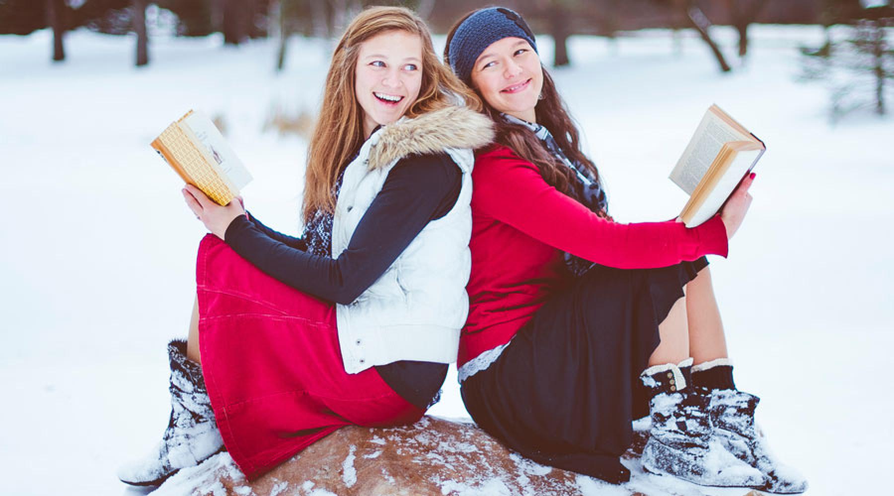 Zwei Mädchen sitzen auf einem Stein in einer tief verschneiten Landschaft, haben Bücher in der Hand und lachen sich an.