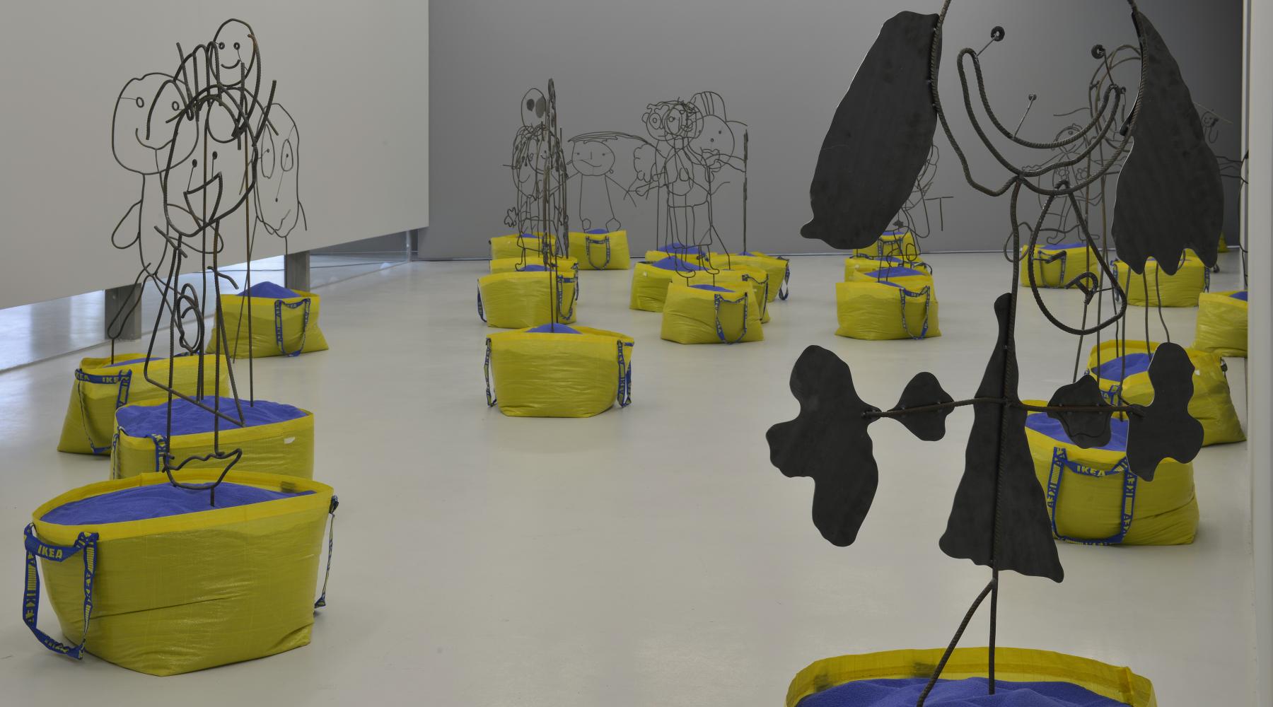 Konstinstallationer står i ett rum på ett galleri. De visar barnsliga figurer i metall som är placerade i gula Ikea-kassar fyllda med blå sand.