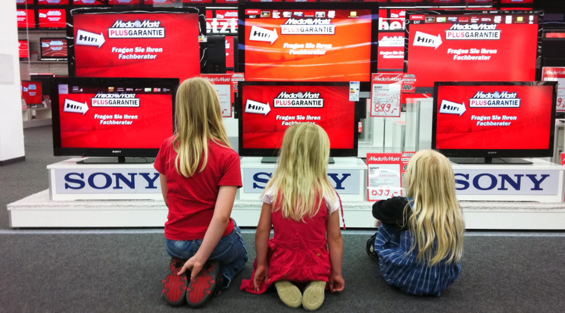 Drei Kinder sitzen vor einer Wand aus Fernsehern in einem Media Markt.