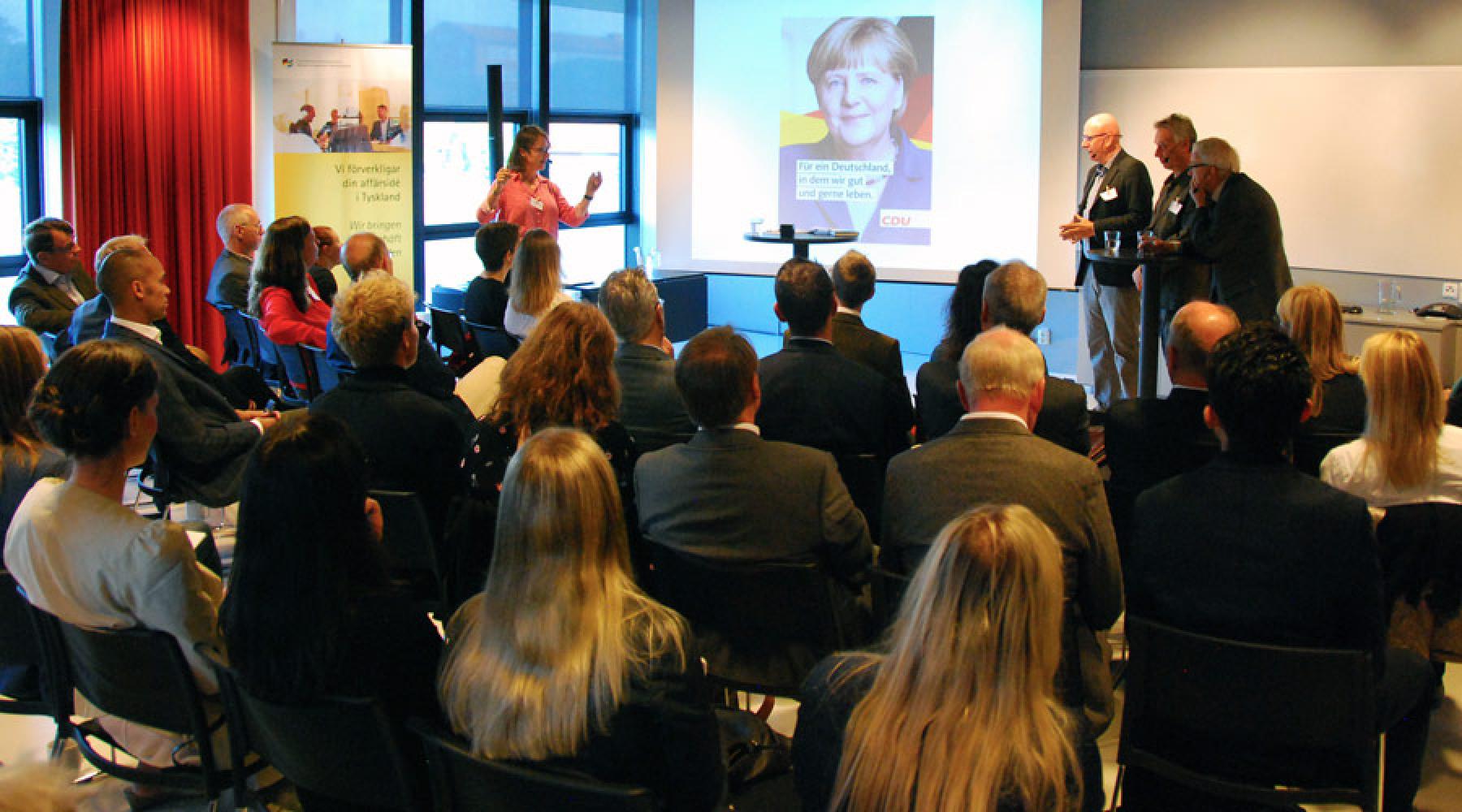 Publiken på Tyskland inför valet-seminariet, längst fram panelen, moderatorn och en powerpoint-bild med en av Angela Merkels valaffischer