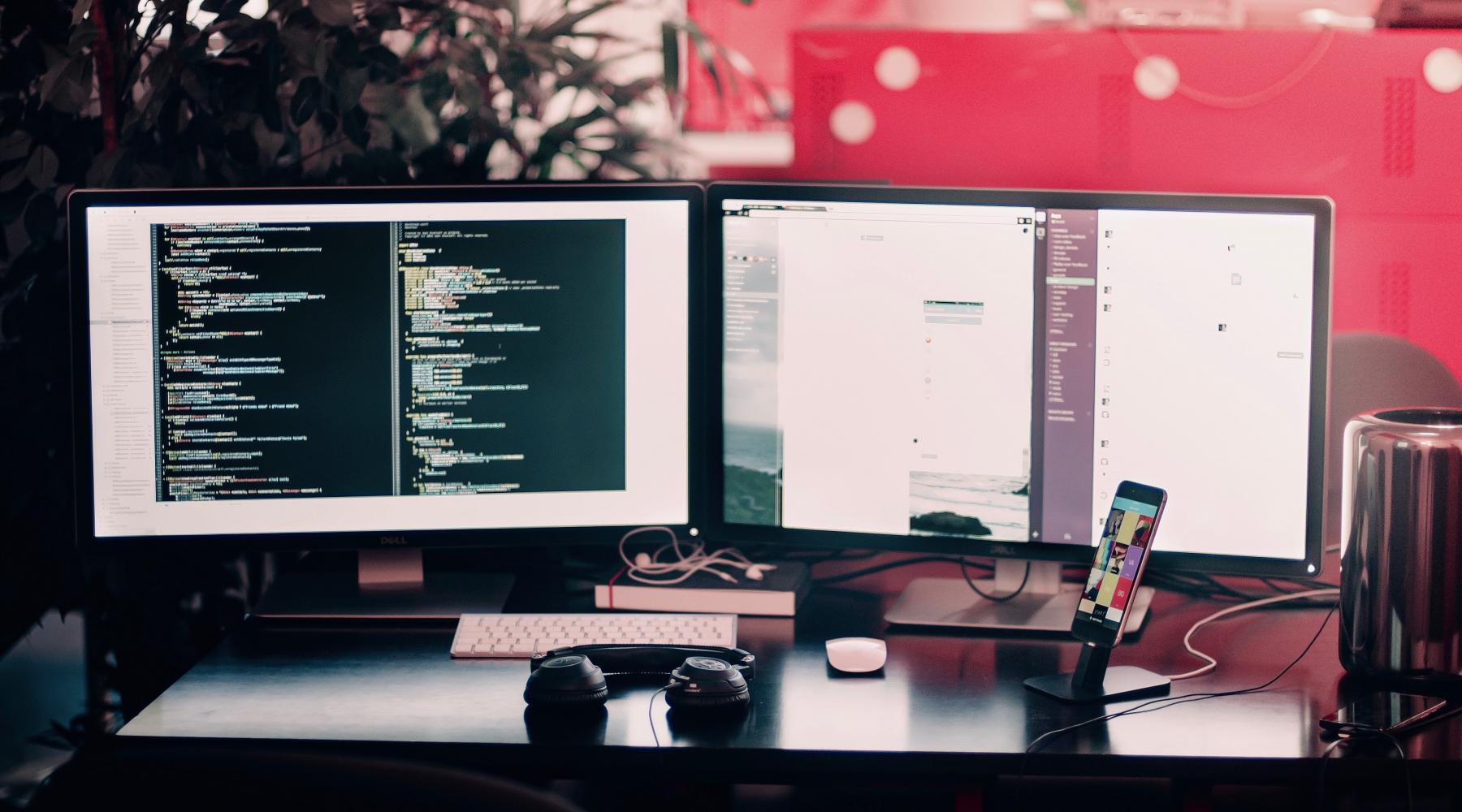 Ein Schreibtisch mit 2 Computer-Bildschirmen. Auf dem linken ist Programmier-Code zu sehen.