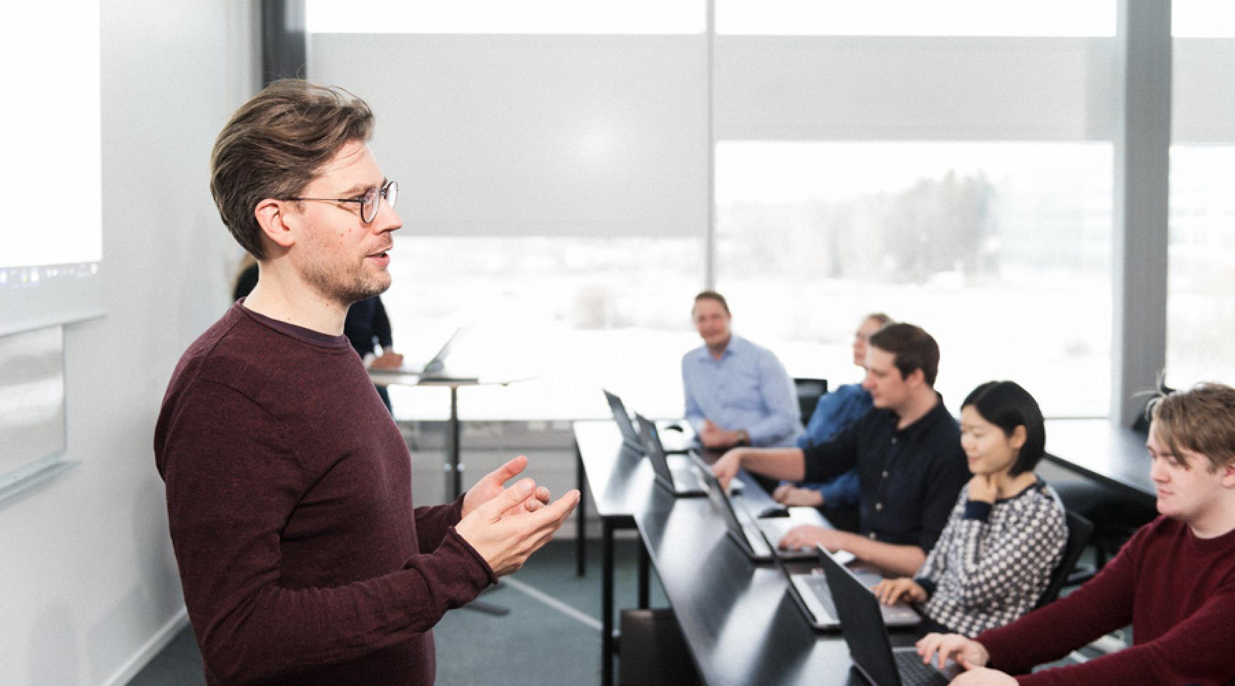 Ein Lehrer unterrichtet eine Gruppe von Erwachsenen, die mit Laptops an Schulbänken sitzen