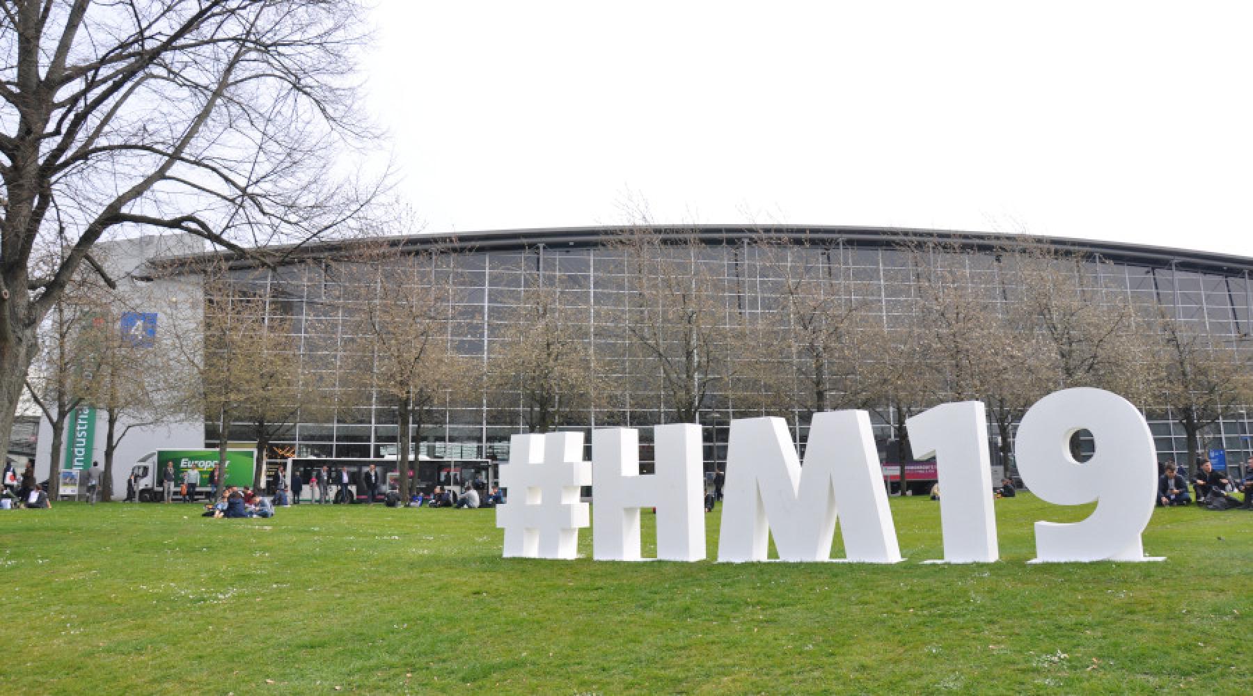 Hannovermässans hashtag #HM19 uppställd på en gräsmatta framför en av mässhallarna