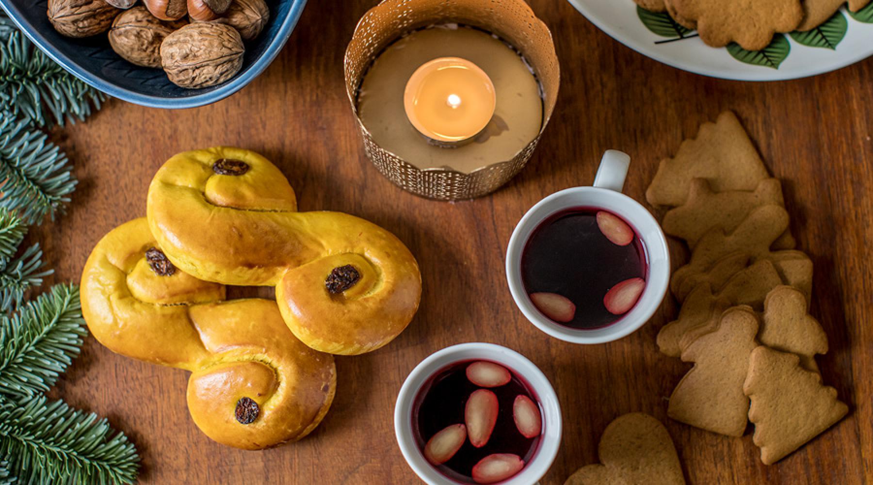 Festligt dukat träbord med ett tänt ljus, två koppar glögg med mandelskivor, pepparkakor i hjärt- och granform, lussekatter med russin, en skål med valnötter och dekorativa grankvistar – en stämningsfull scen för ett svenskt julmingel.