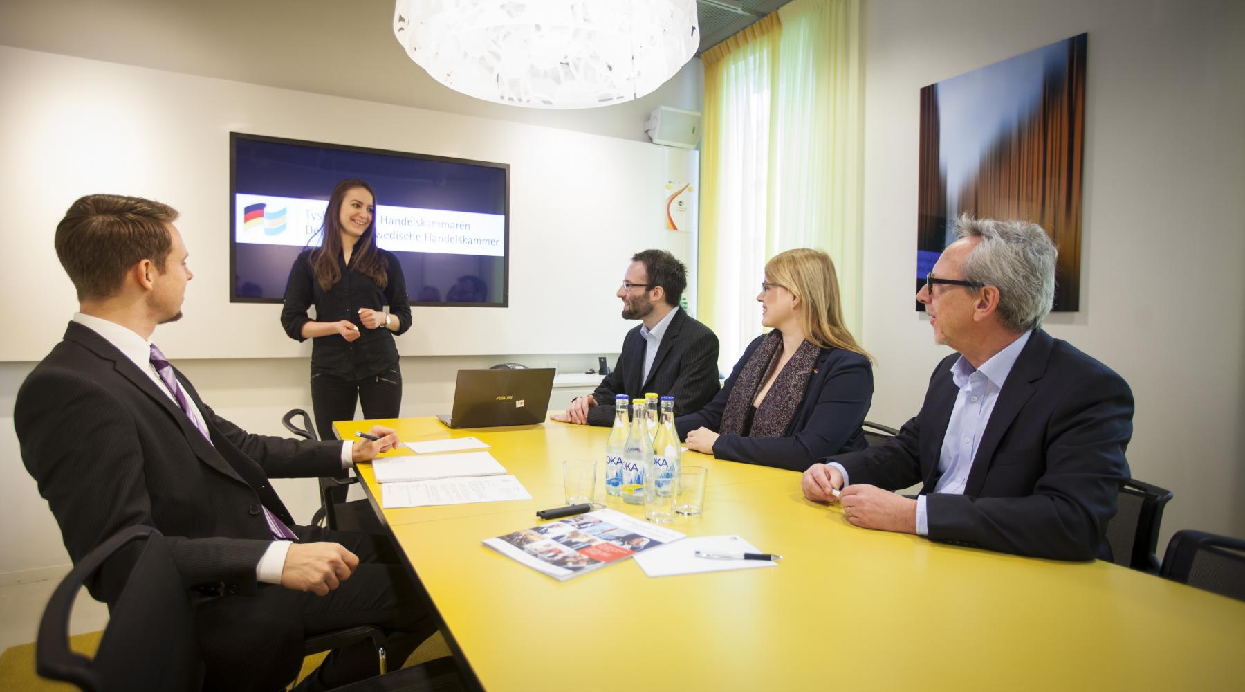 En kvinna står framför en grupp människor som sitter runt ett konferensbord.